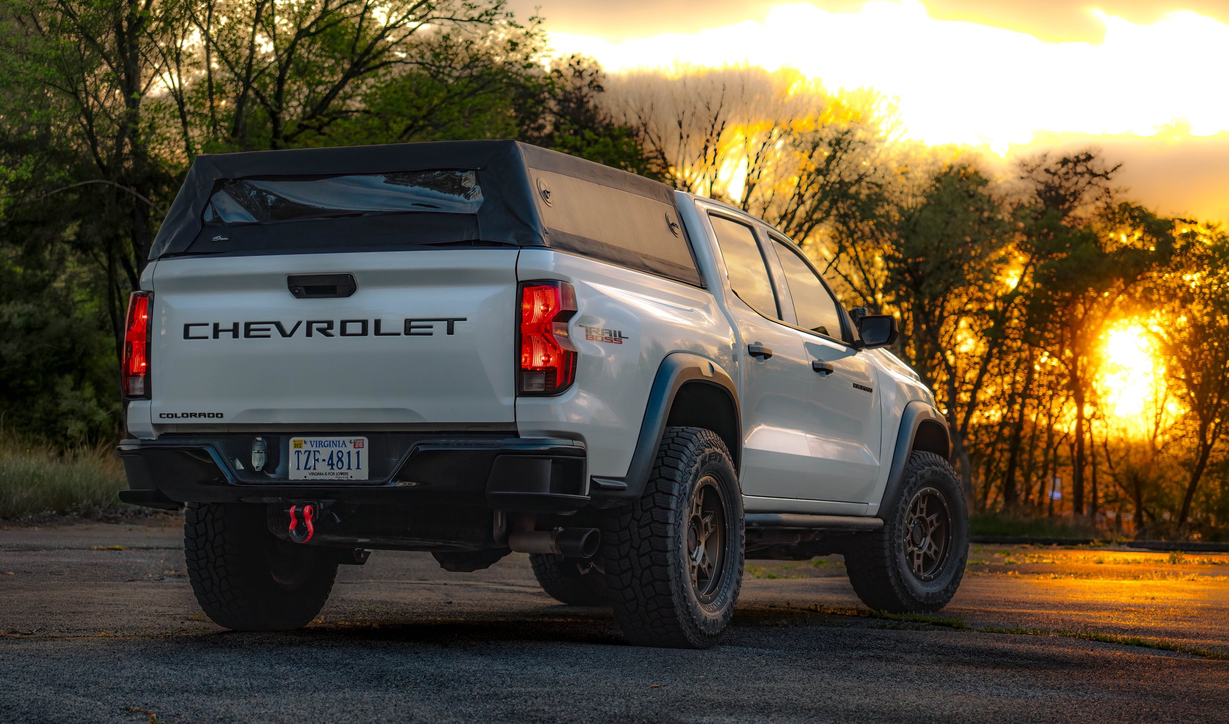 A white Chevrolet pickup truck with a camper shell is parked on a paved surface during sunset, with trees in the background.
