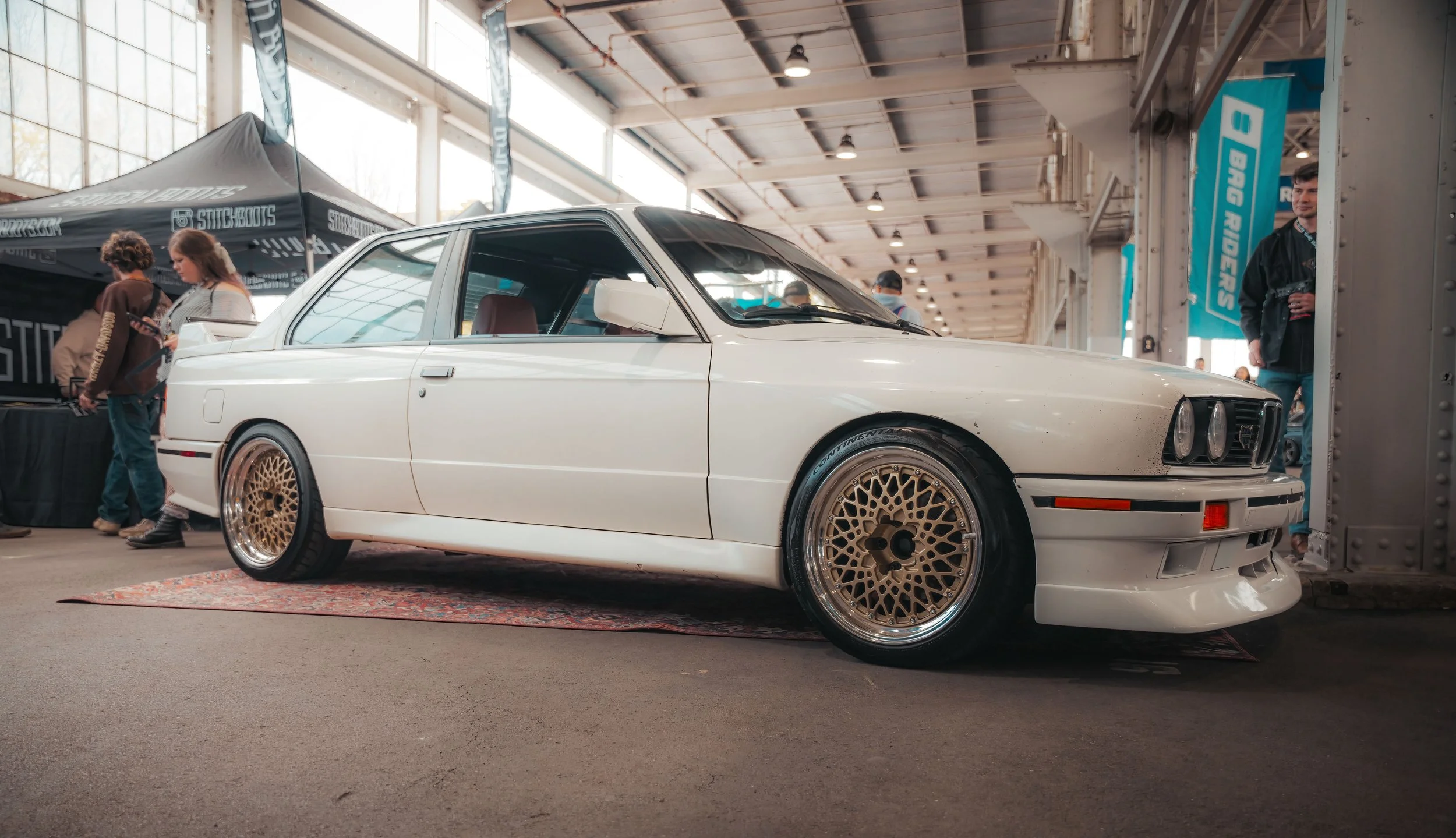 A white vintage sports car with gold mesh wheels displayed indoors at a car show, with people walking around and booths in the background.
