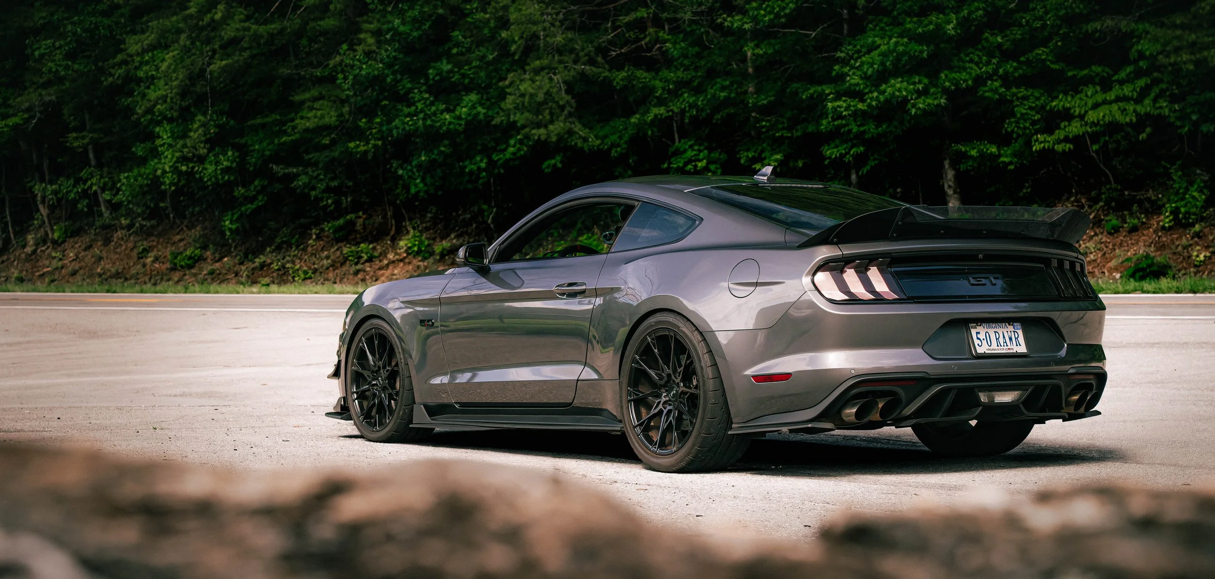 A Carbon Gray Mustang GT with a spoiler, black wheels, and dual exhaust pipes, parked on the side of a road with a forested background.