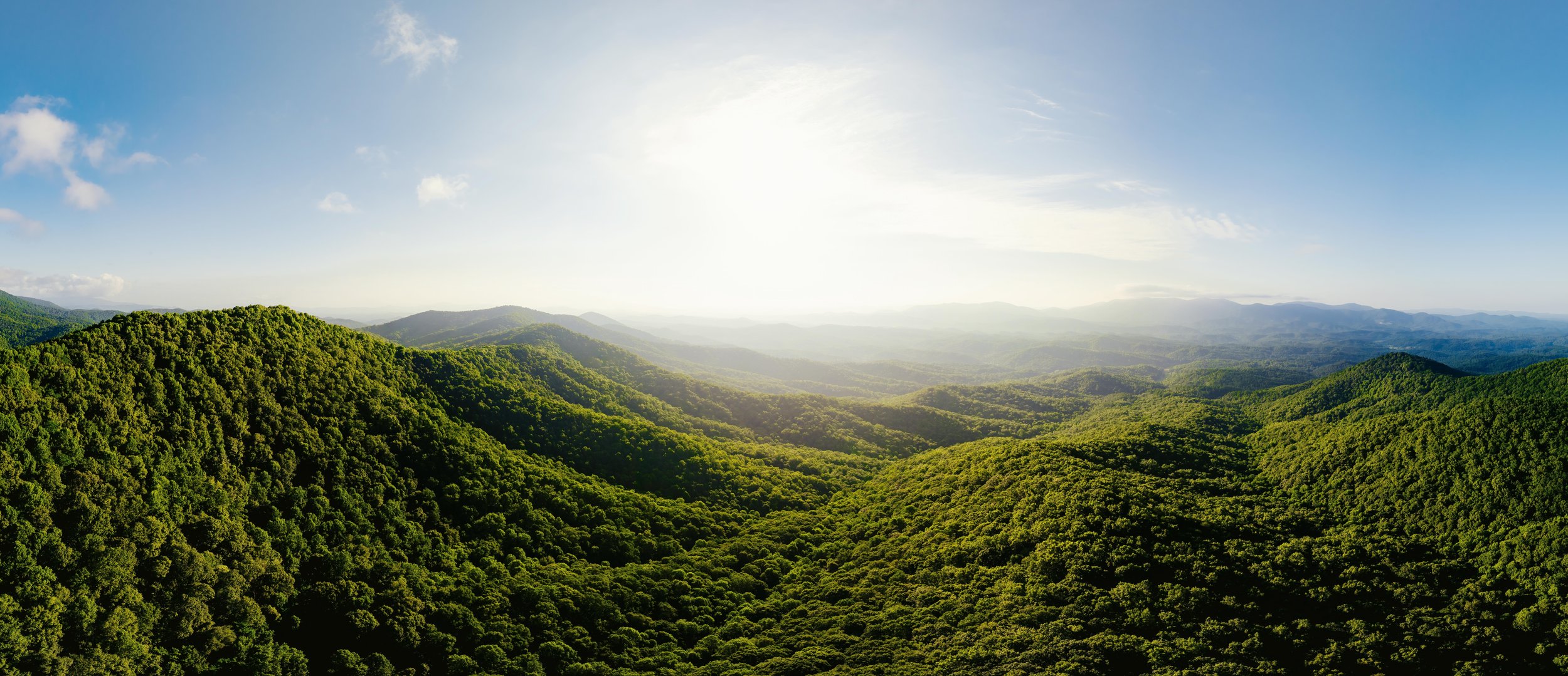 A panoramic view of green forested hills and mountains under a bright blue sky with some clouds and the sun shining.