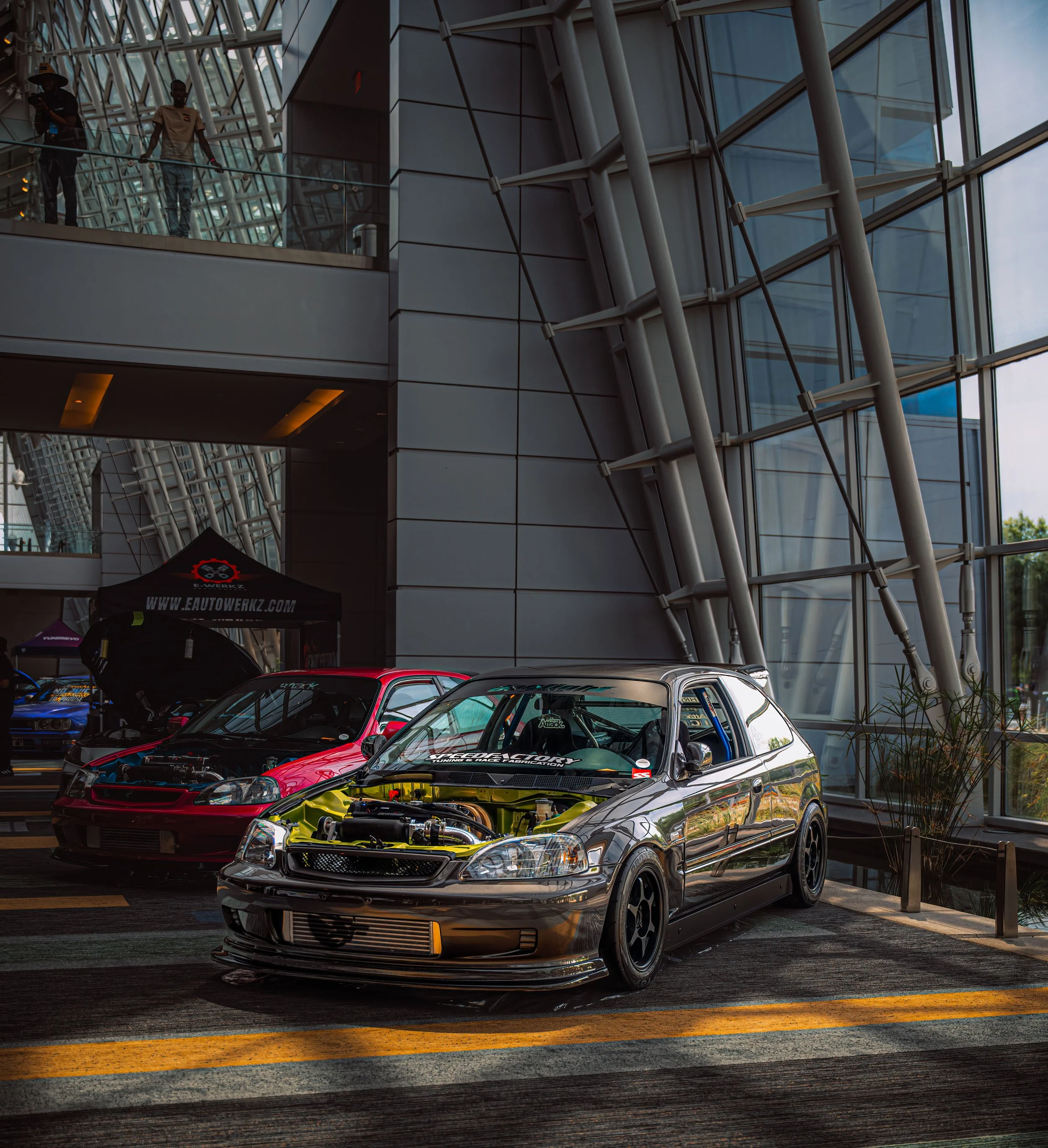 A silver modified Honda Civic hatchback with its hood open, showing the engine, on display at an indoor car show. Behind it, there is a red car and other vehicles, with a black tent that has the website 'Eautowrkz.com' printed on it.