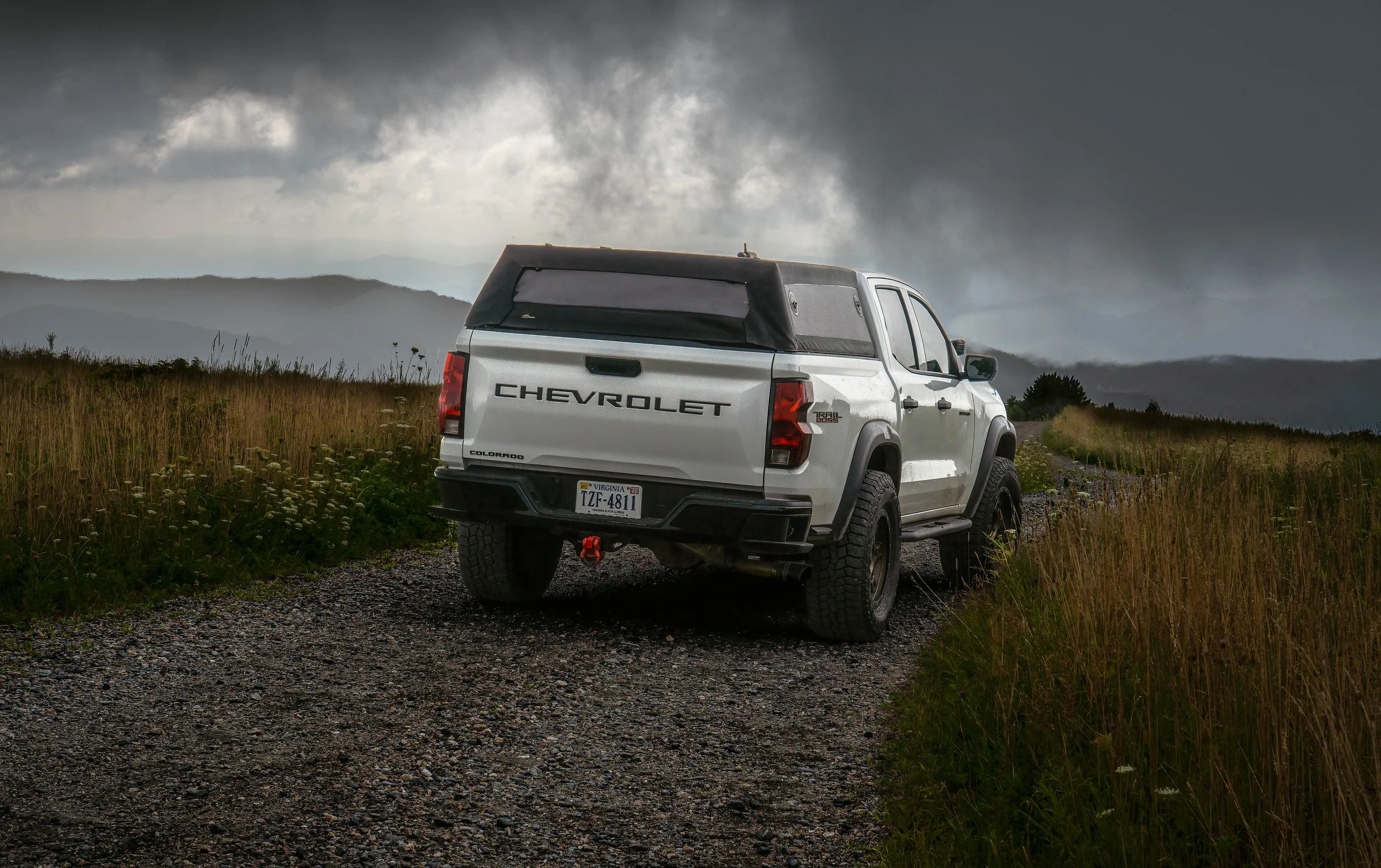 A white Chevrolet Colorado pickup truck with a black camper shell driving on a dirt trail in a grassy field under dark stormy sky.