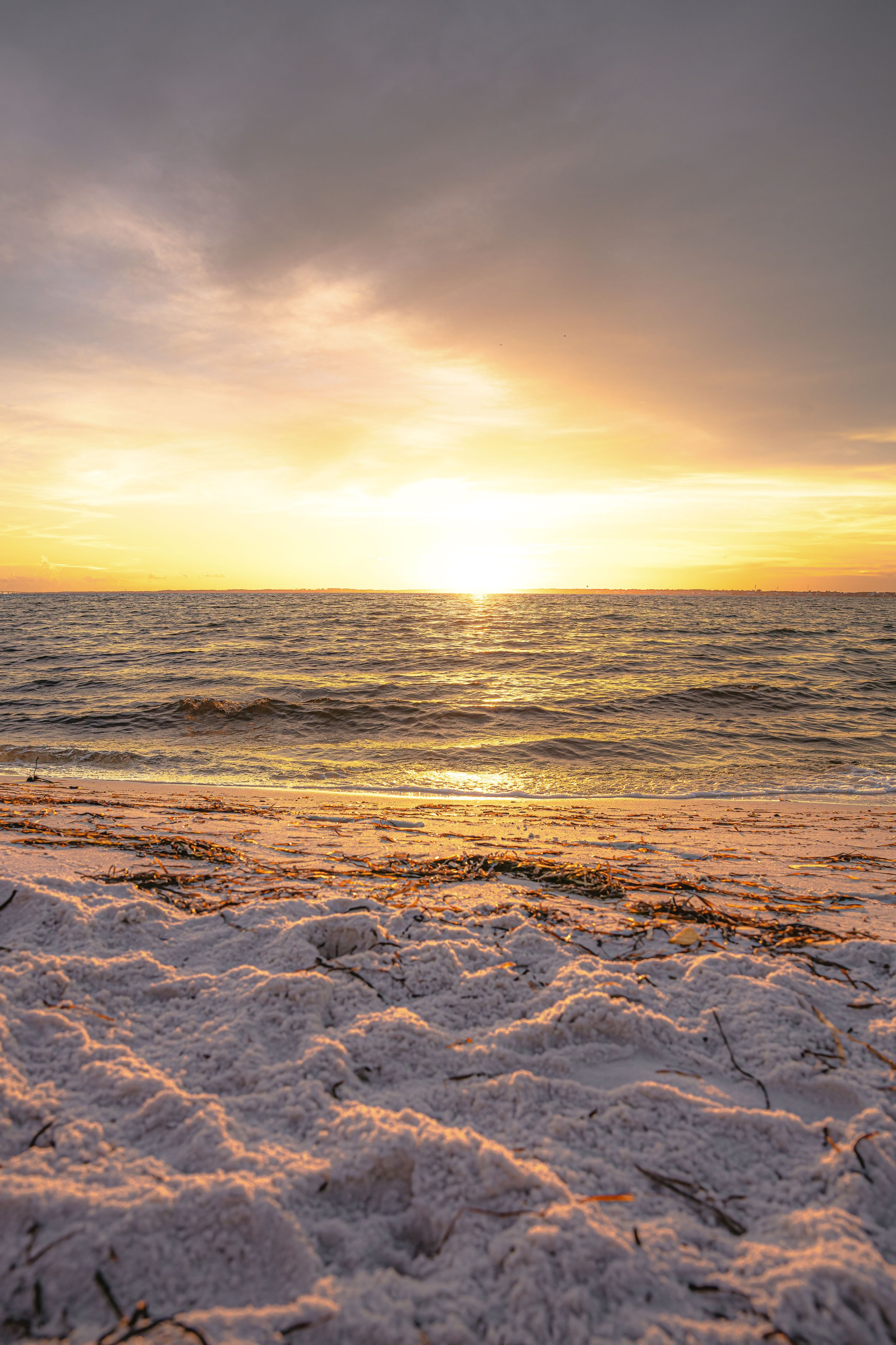 Sunset over the ocean with a sandy beach in the foreground, some seaweed and debris scattered on the sand.