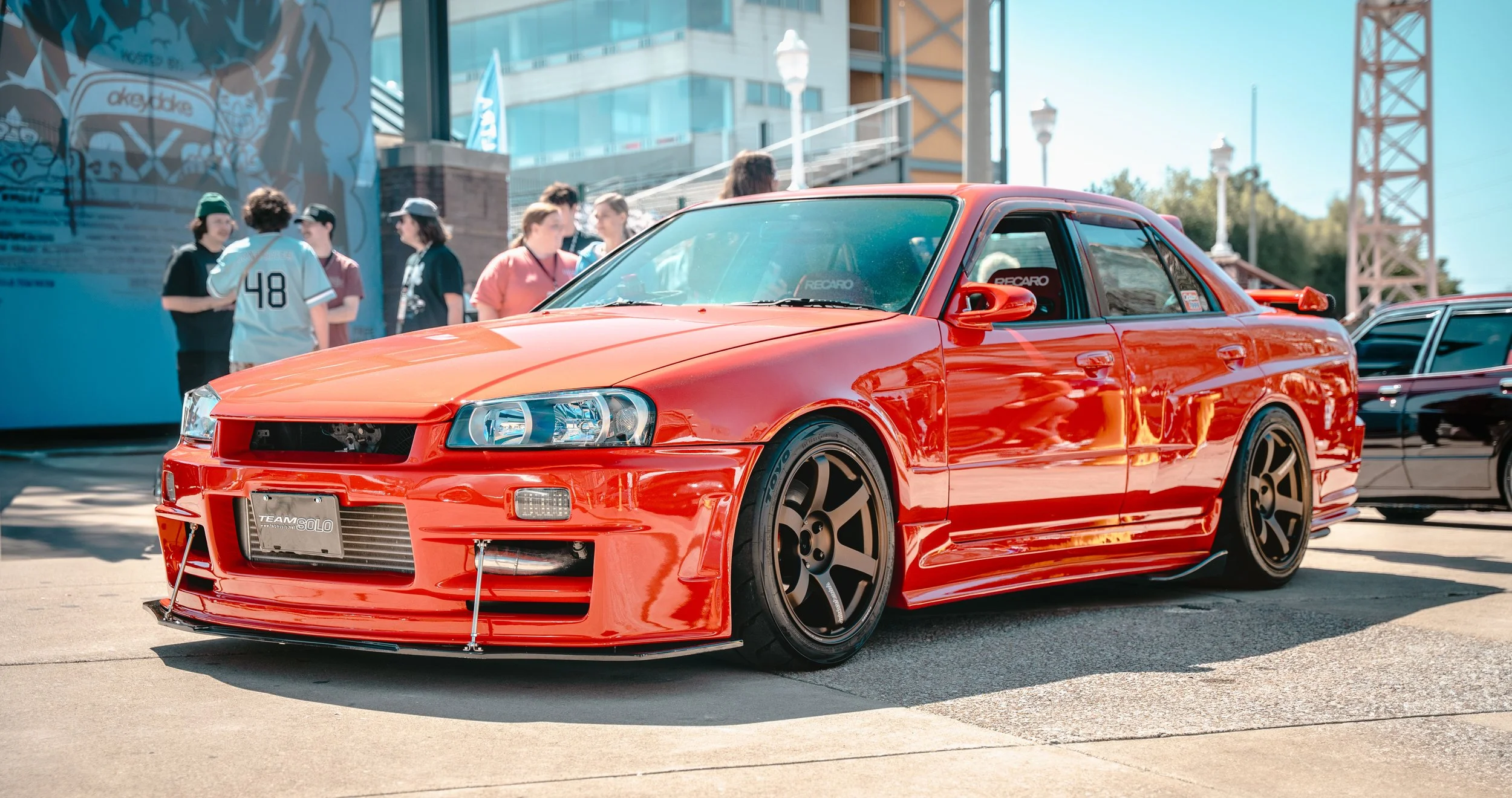 Red modified Nissan Skyline GT-R R34 sports car at a car show with people in the background.