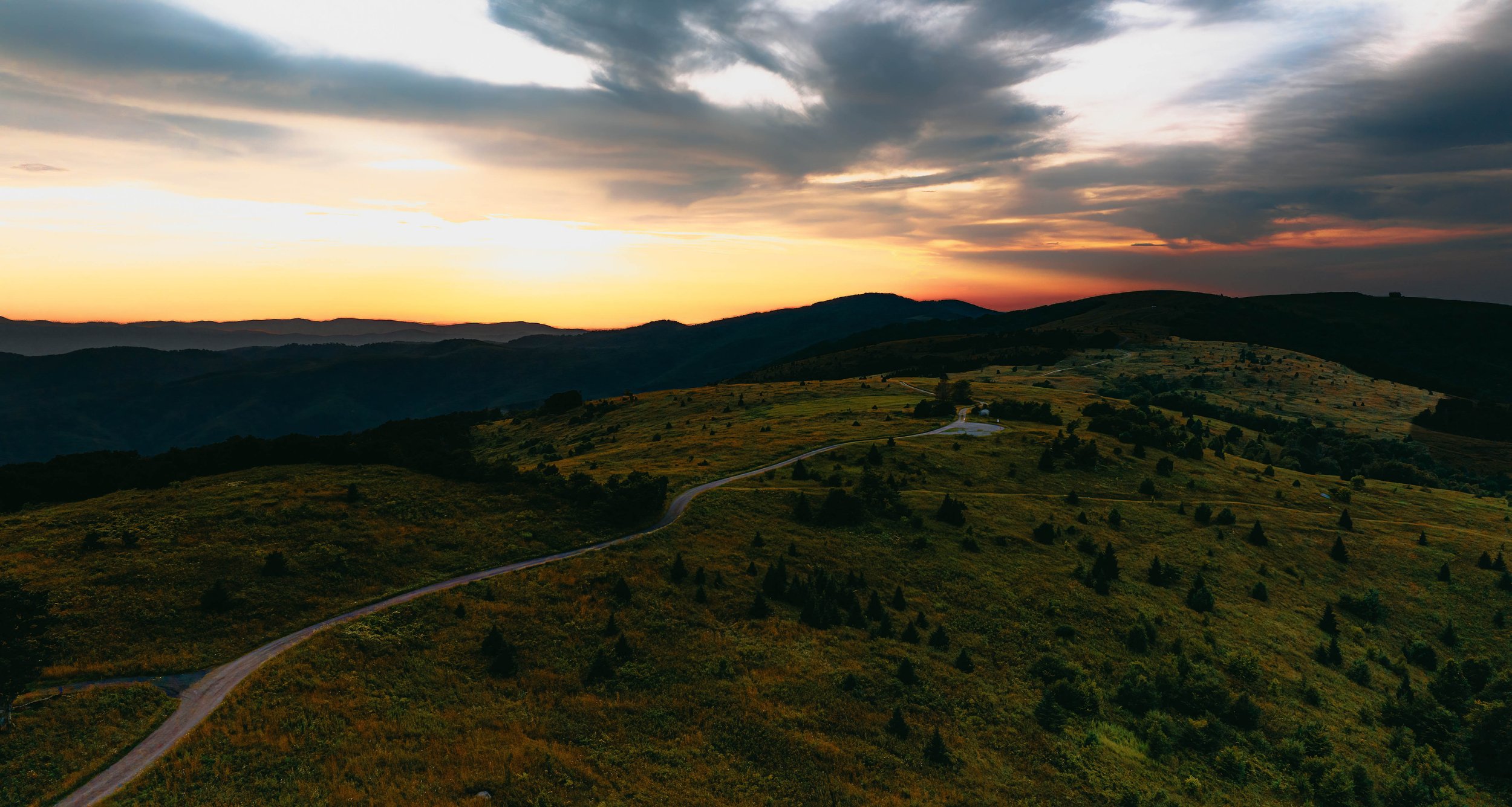 Scenic view of rolling hills at sunset with a winding road and dark clouds in the sky.