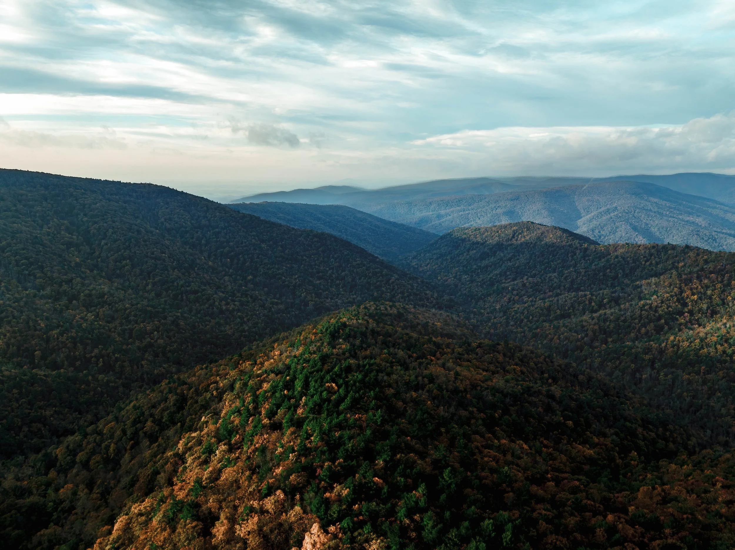 Expansive view of rolling mountains covered in dense forests with a cloudy sky above.