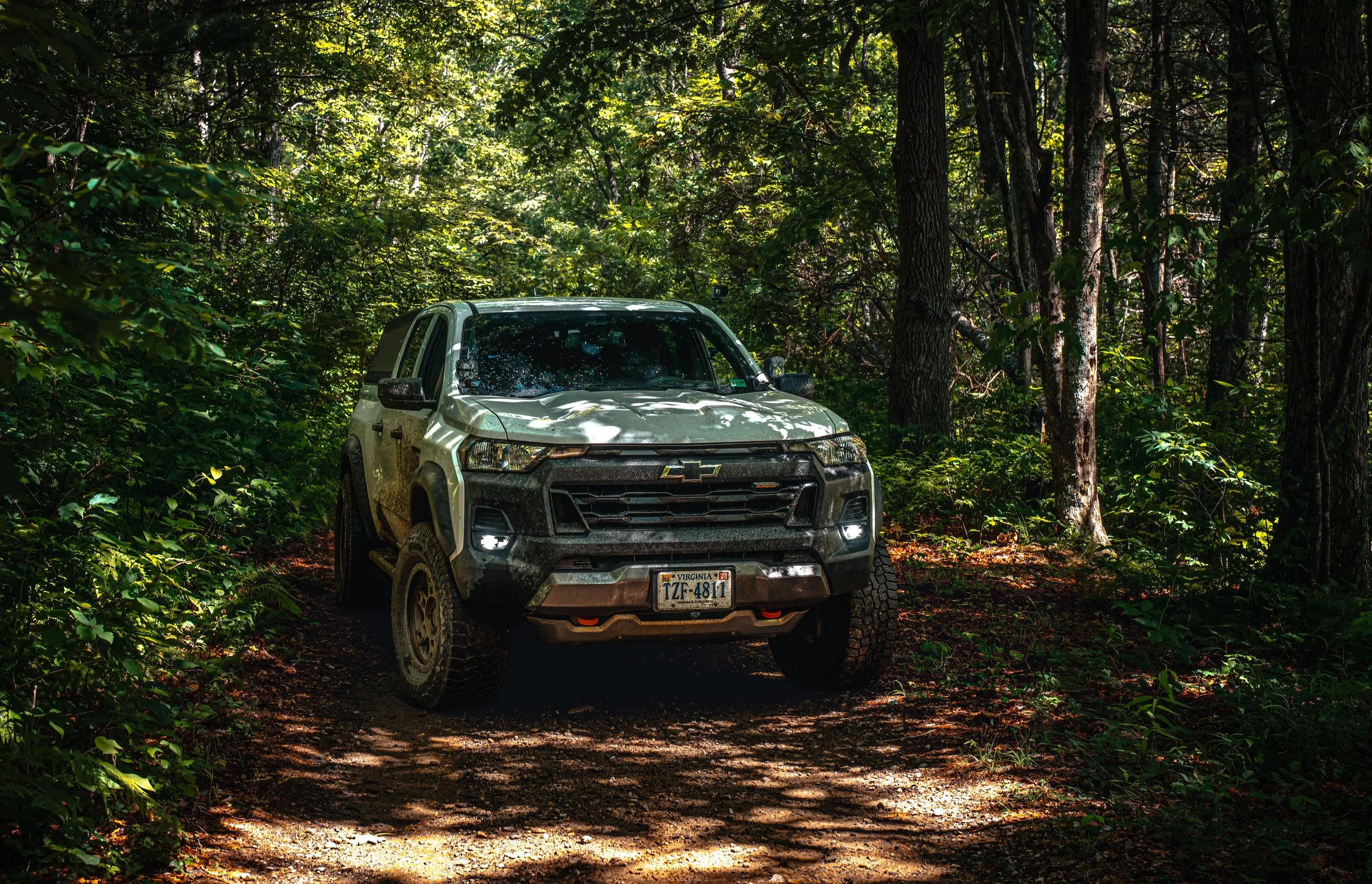 A white off-road vehicle with muddy tires parked on a dirt trail in a dense green forest, with sunlight filtering through the trees.