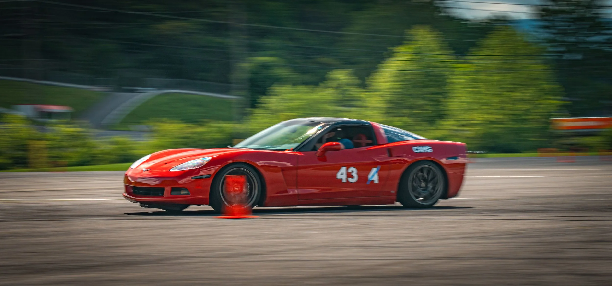 Red sports car with the number 43 racing on an outdoor track, with motion blur and green trees in the background.