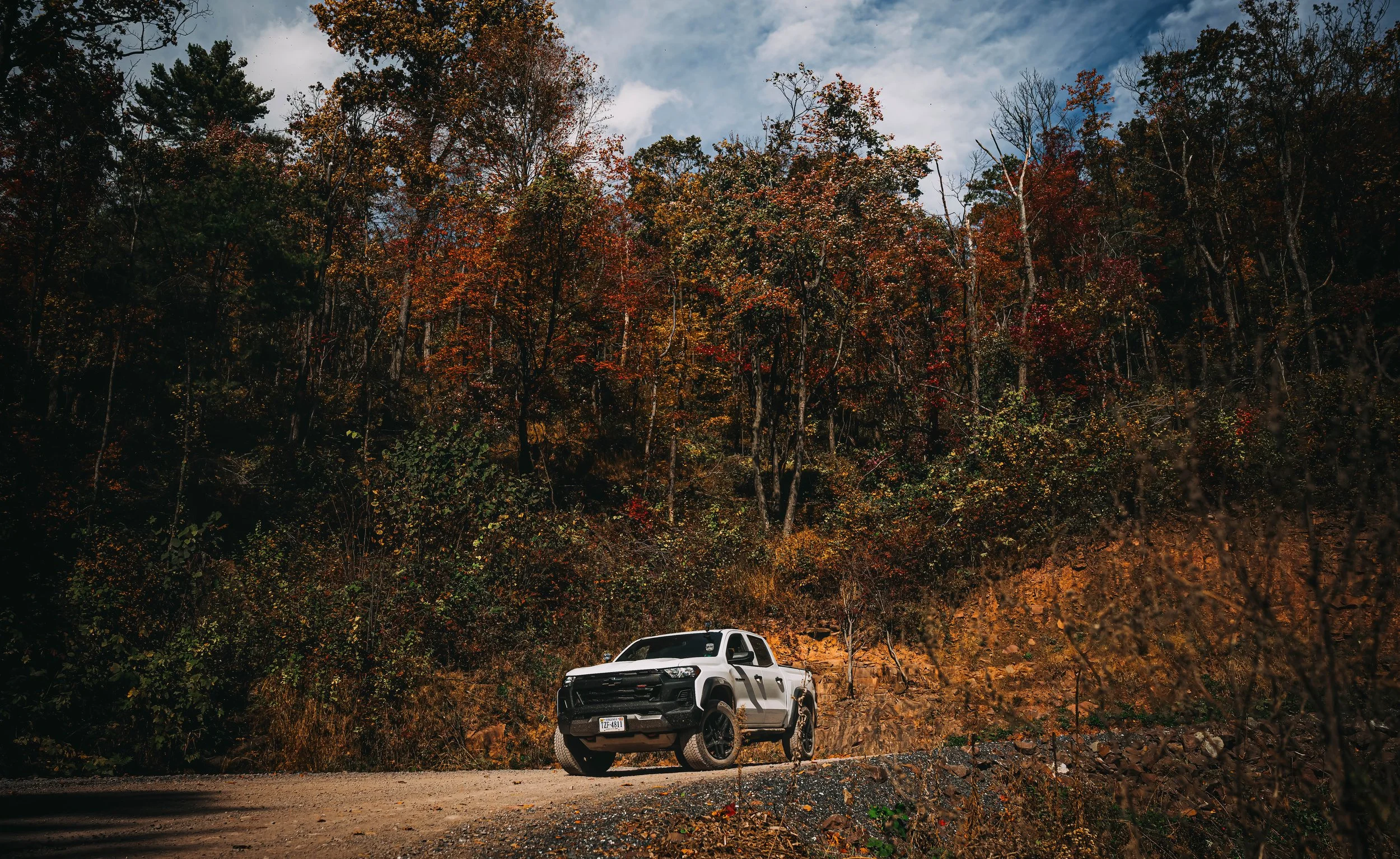 A black and white pickup truck parked on a dirt road in front of a forested hillside with autumn-colored trees under a partly cloudy sky.