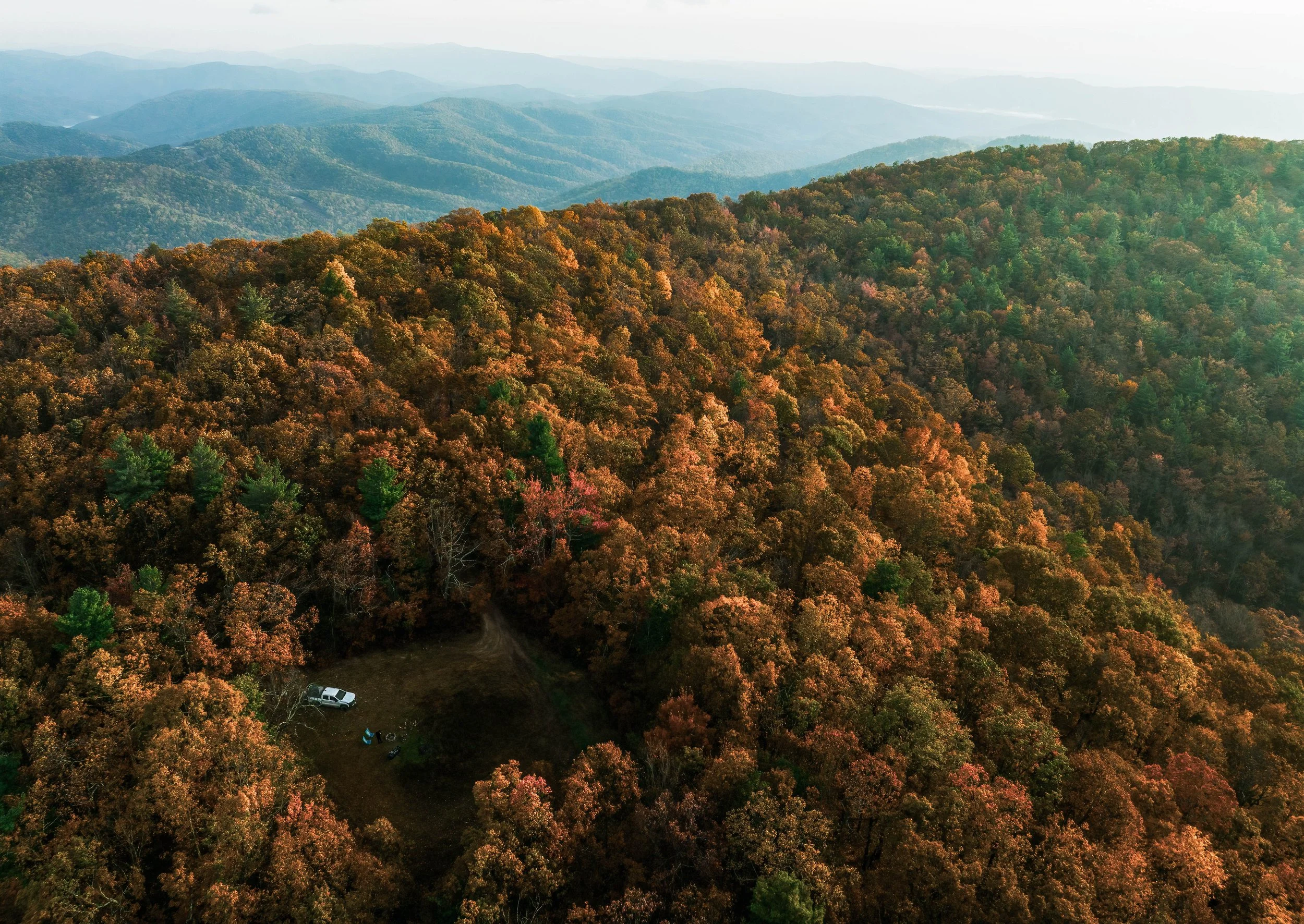 Aerial view of a dense forested mountain landscape with fall foliage in various shades of orange and green, and a small clearing with a white vehicle and camping gear.
