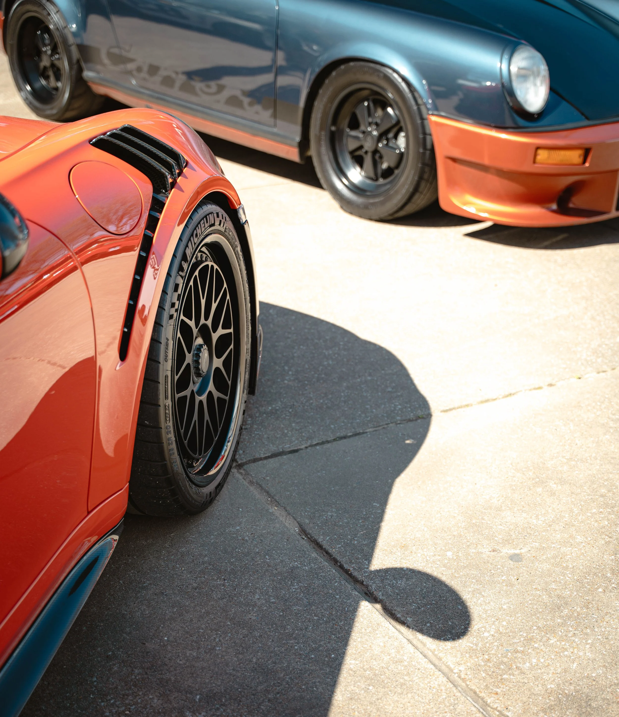 Close-up of a red sports car with black wheels and aerodynamic vents on the fender, parked on a driveway; a blue sports car with tinted windows and black rims behind it.