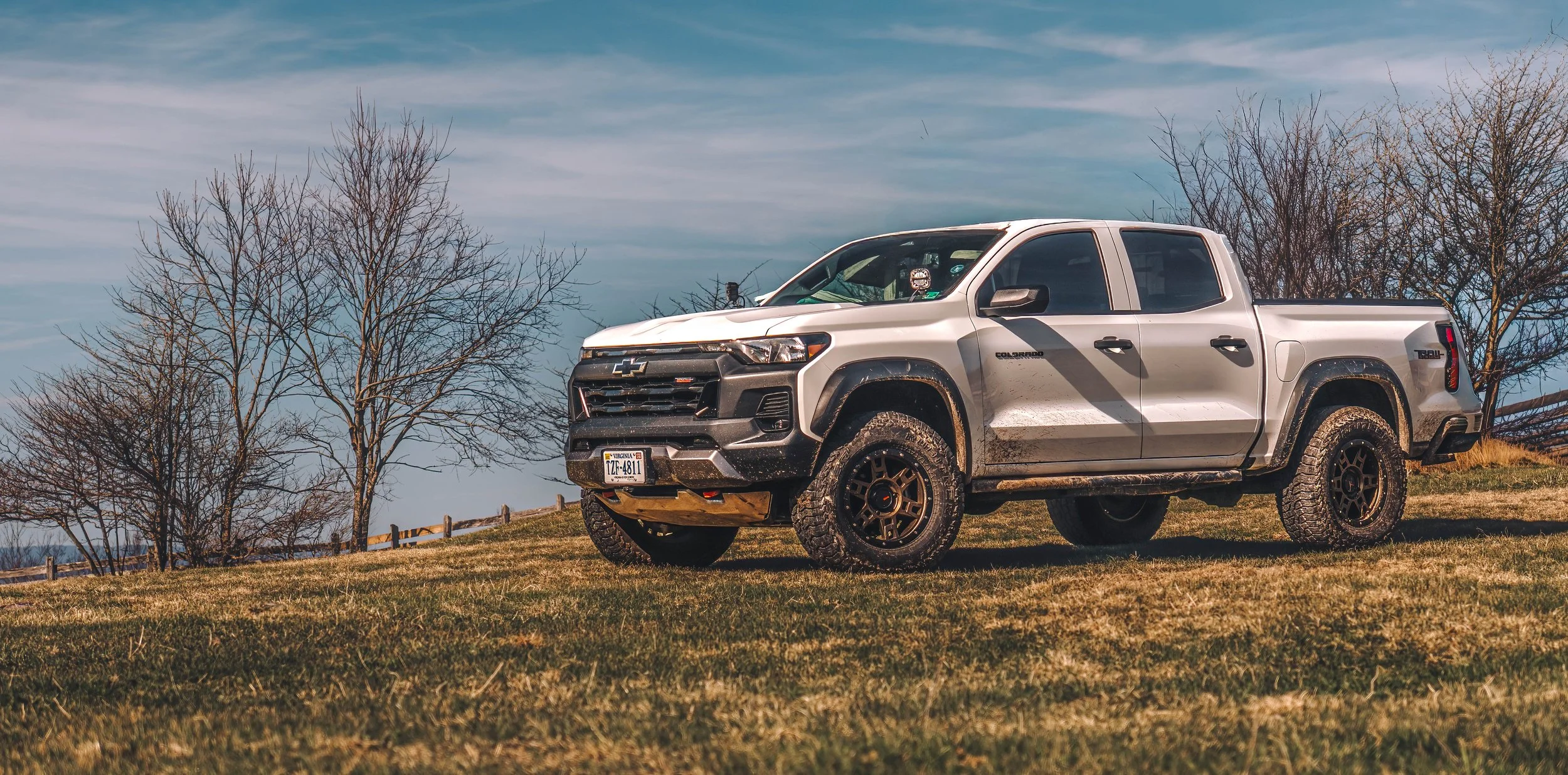 A white Chevrolet Colorado pickup truck with off-road tires parked on a grassy hill with leafless trees and a cloudy sky in the background.