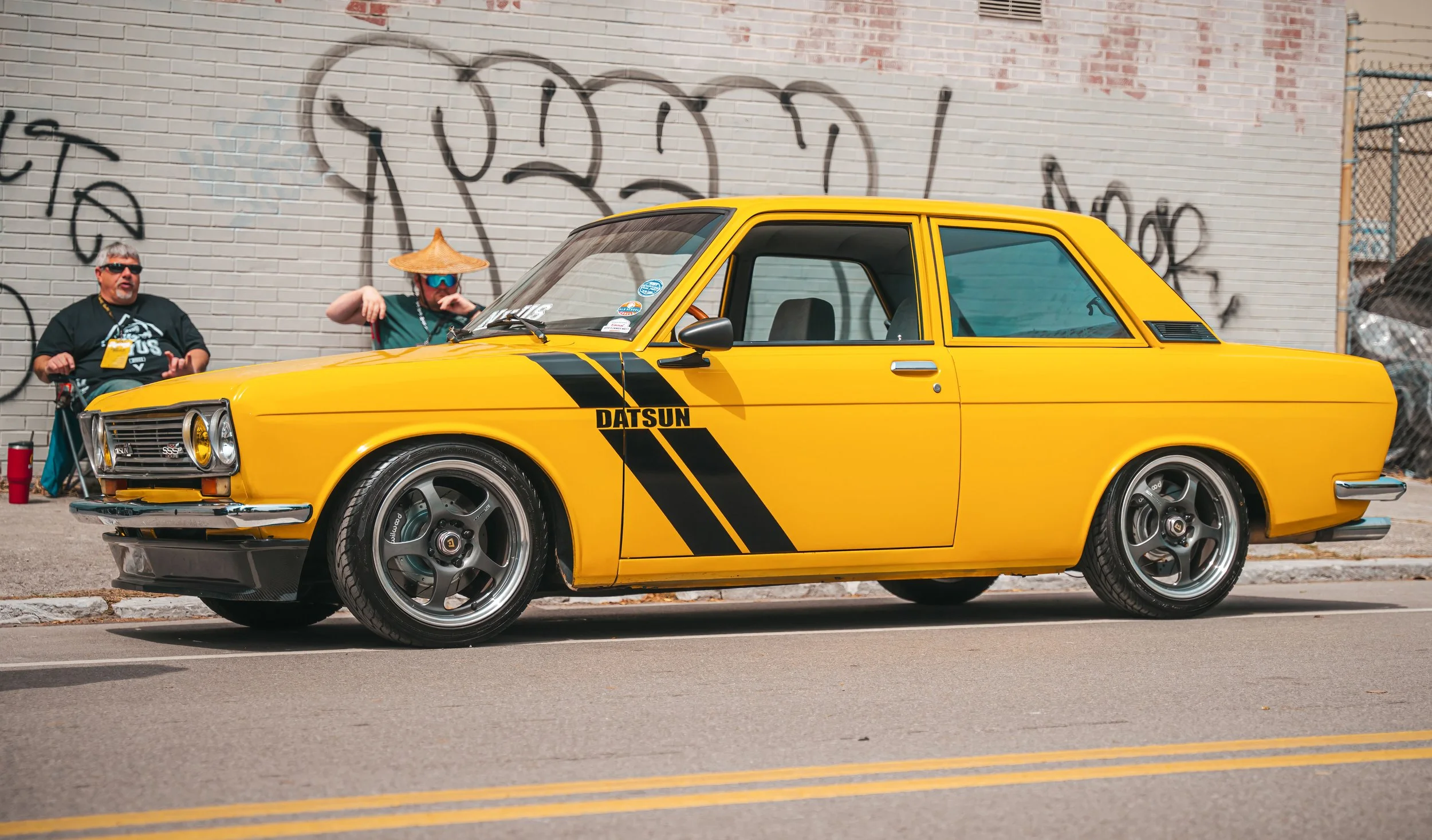 A yellow vintage Datsun car with black racing stripes parked on the street with two men sitting in the background near a graffiti-covered brick wall.