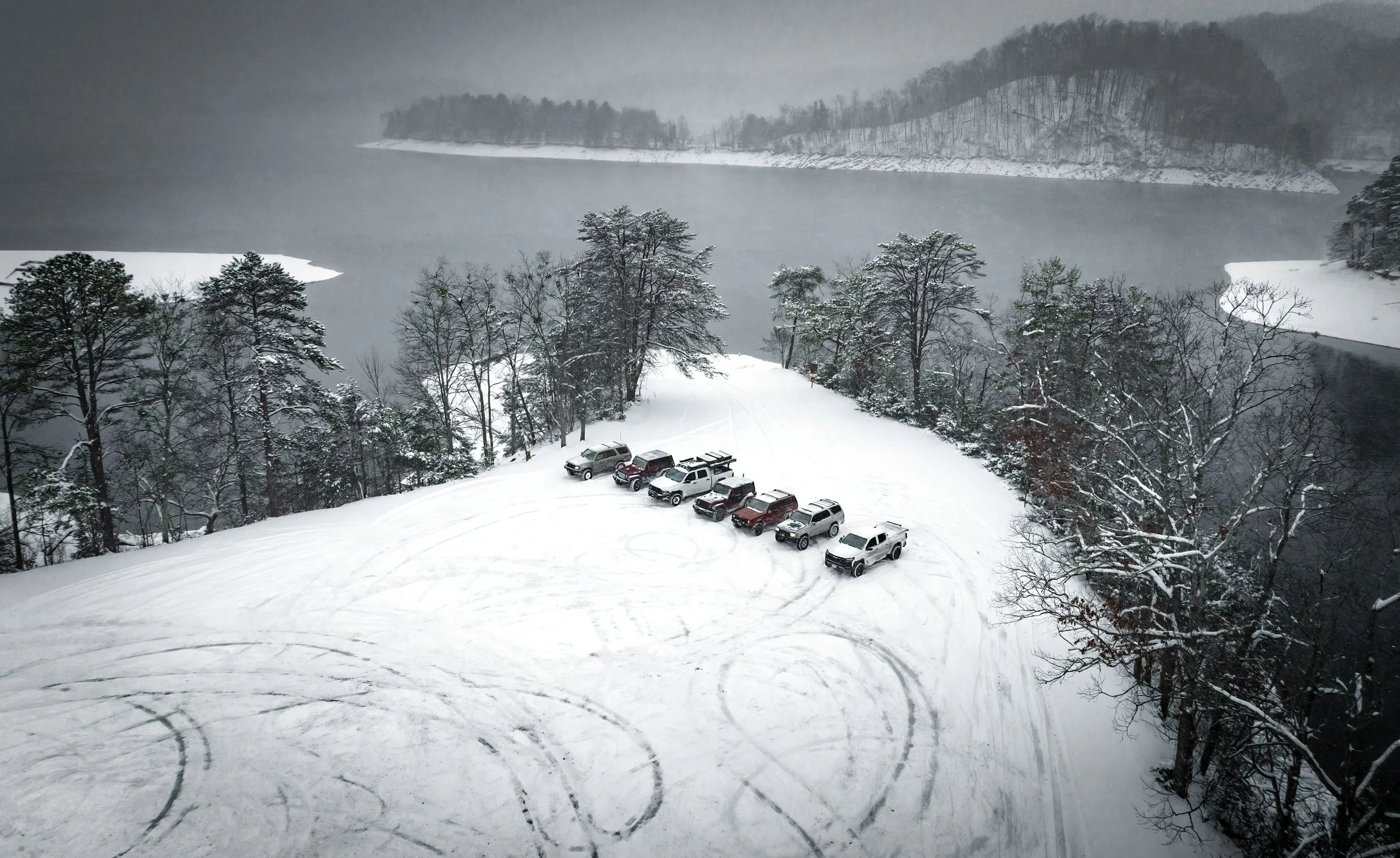 Snow-covered parking lot with multiple vehicles parked near a lake surrounded by snow and trees.
