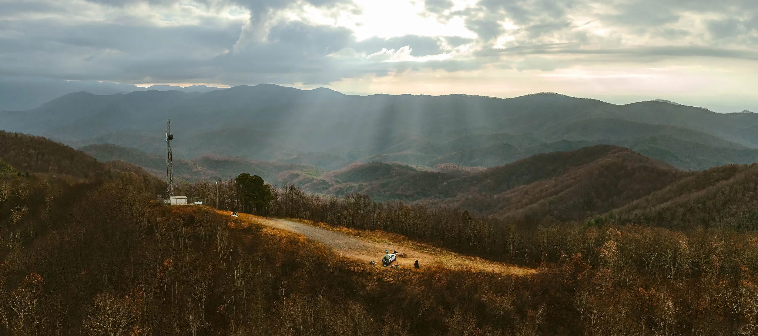 A mountain landscape with rolling hills and a cloudy sky. There is a radio tower on a hill and a Chevy Truck parked on a dirt clearing surrounded by trees.
