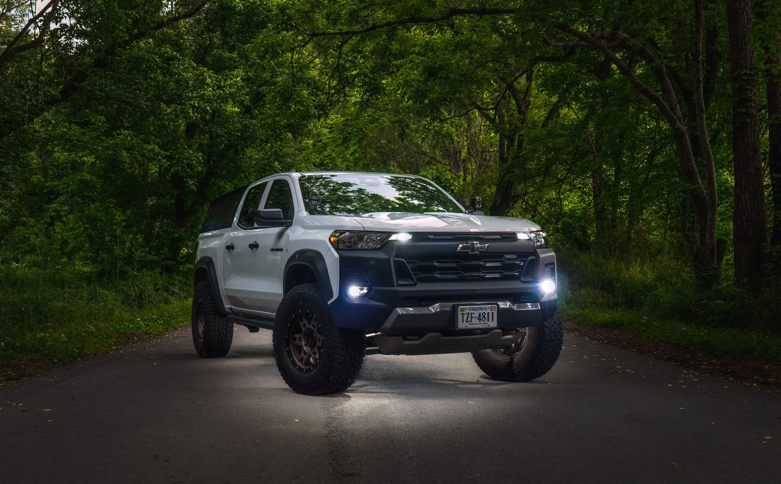 A white Chevrolet SUV parked on a paved road surrounded by lush green trees in a forested area.