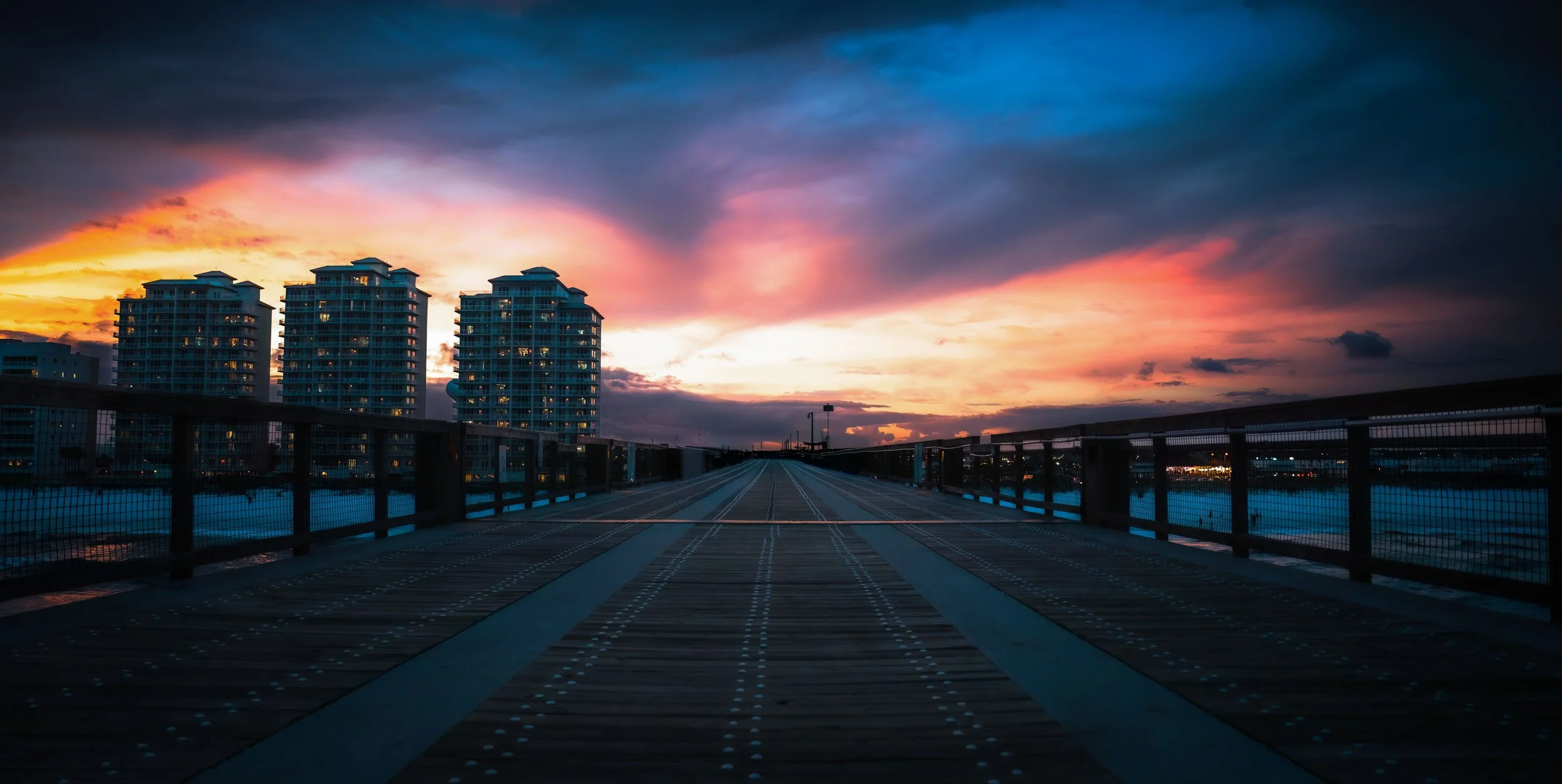 Empty wooden pier or bridge with metal railings over water, with high-rise buildings in the background, under a dramatic colorful sky at sunset.
