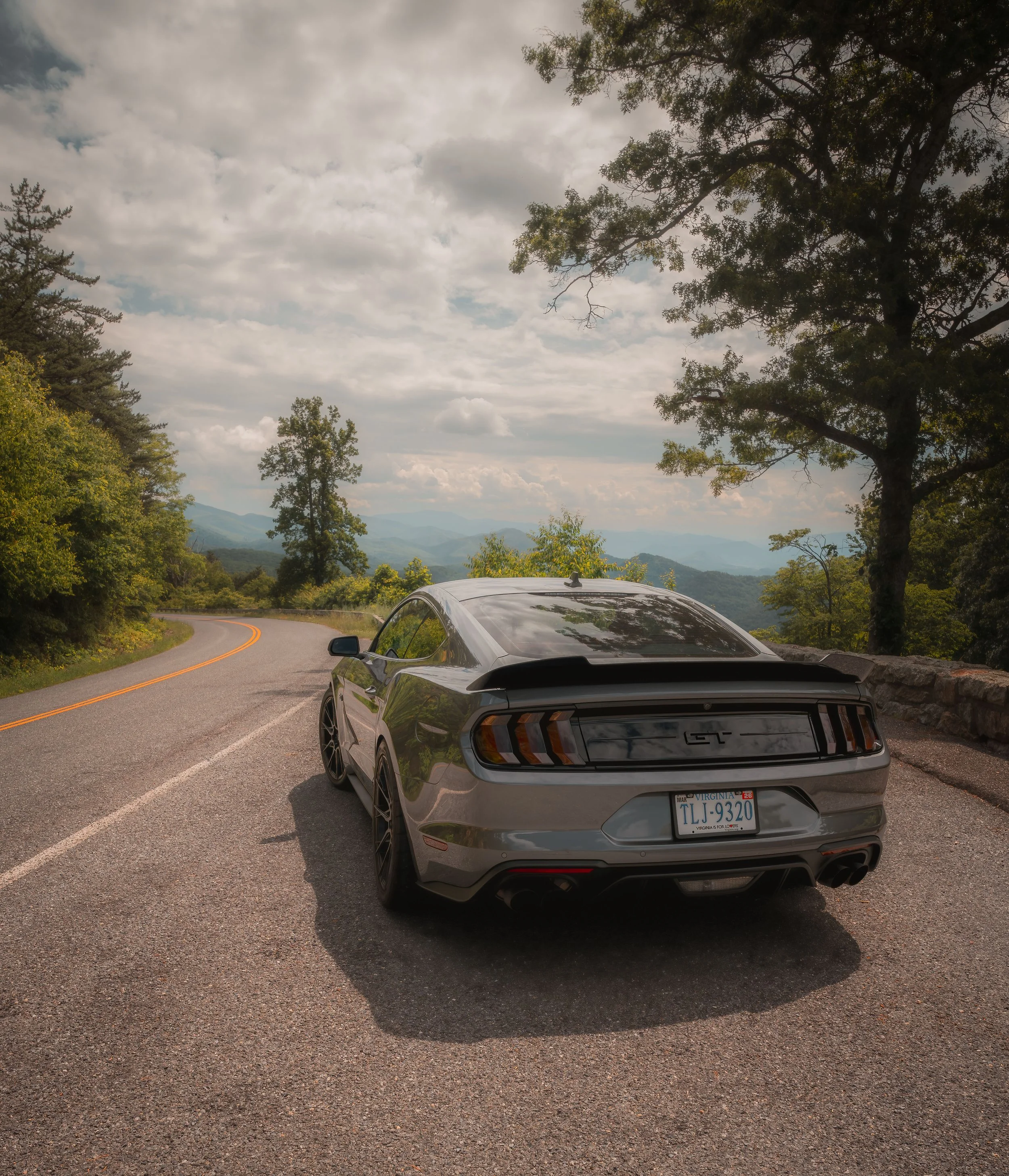 A Gray sports car with a Virginia license plate parked on a winding mountain road surrounded by trees and overlooking a scenic landscape with mountains in the distance under a cloudy sky.