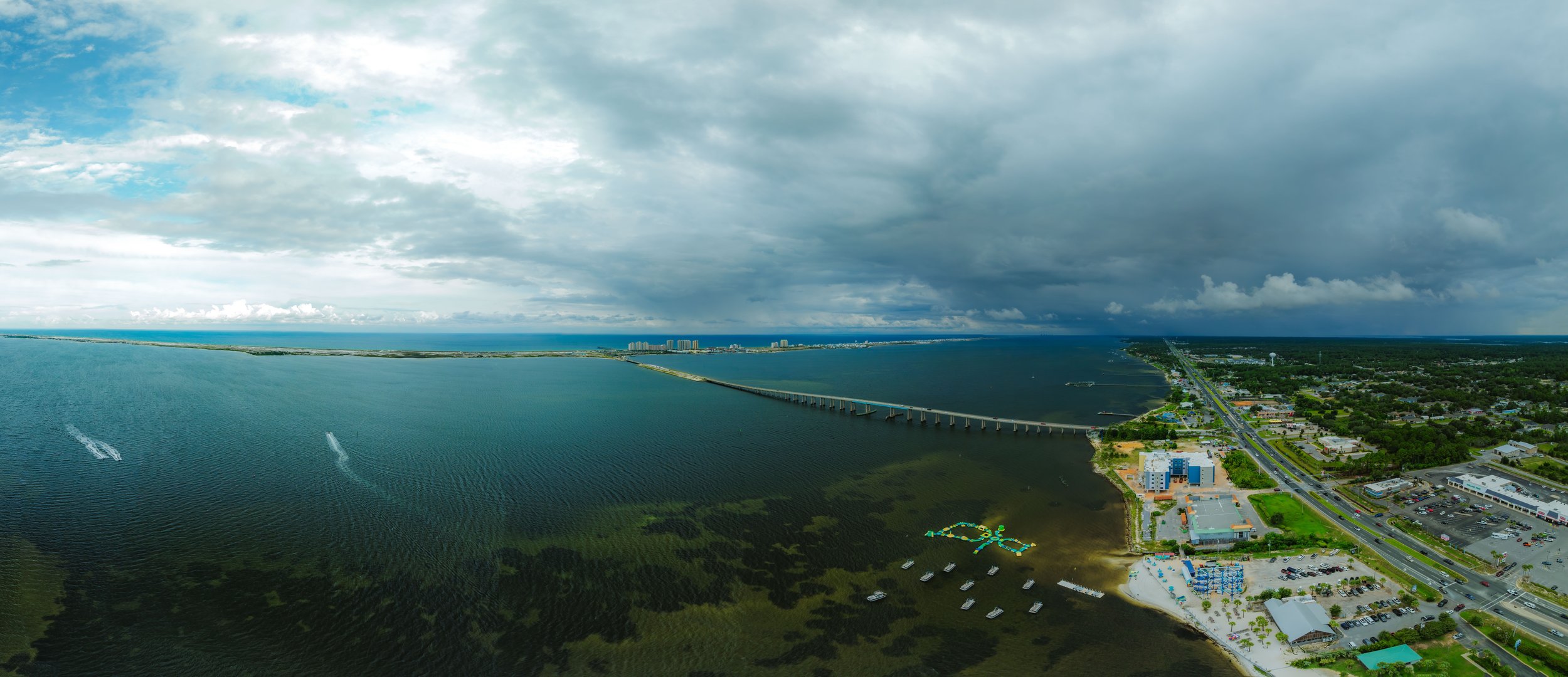 Aerial view of a coastal city with a bridge crossing a large body of water, with boats and water activities, next to a commercial area with parking lots and buildings, under a cloudy sky.