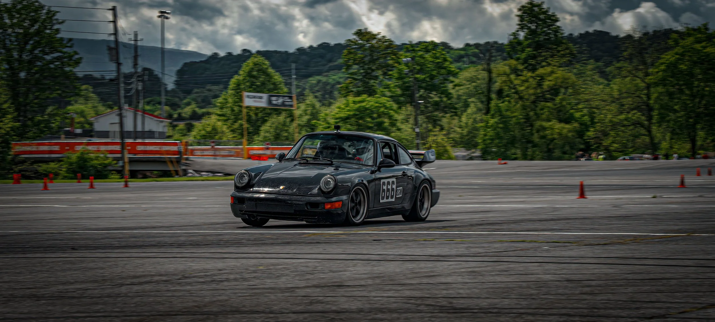 A black Porsche 911 race car with the number 666 on the side, driving on a track with orange cones, surrounded by green trees and a mountainous backdrop under a cloudy sky.