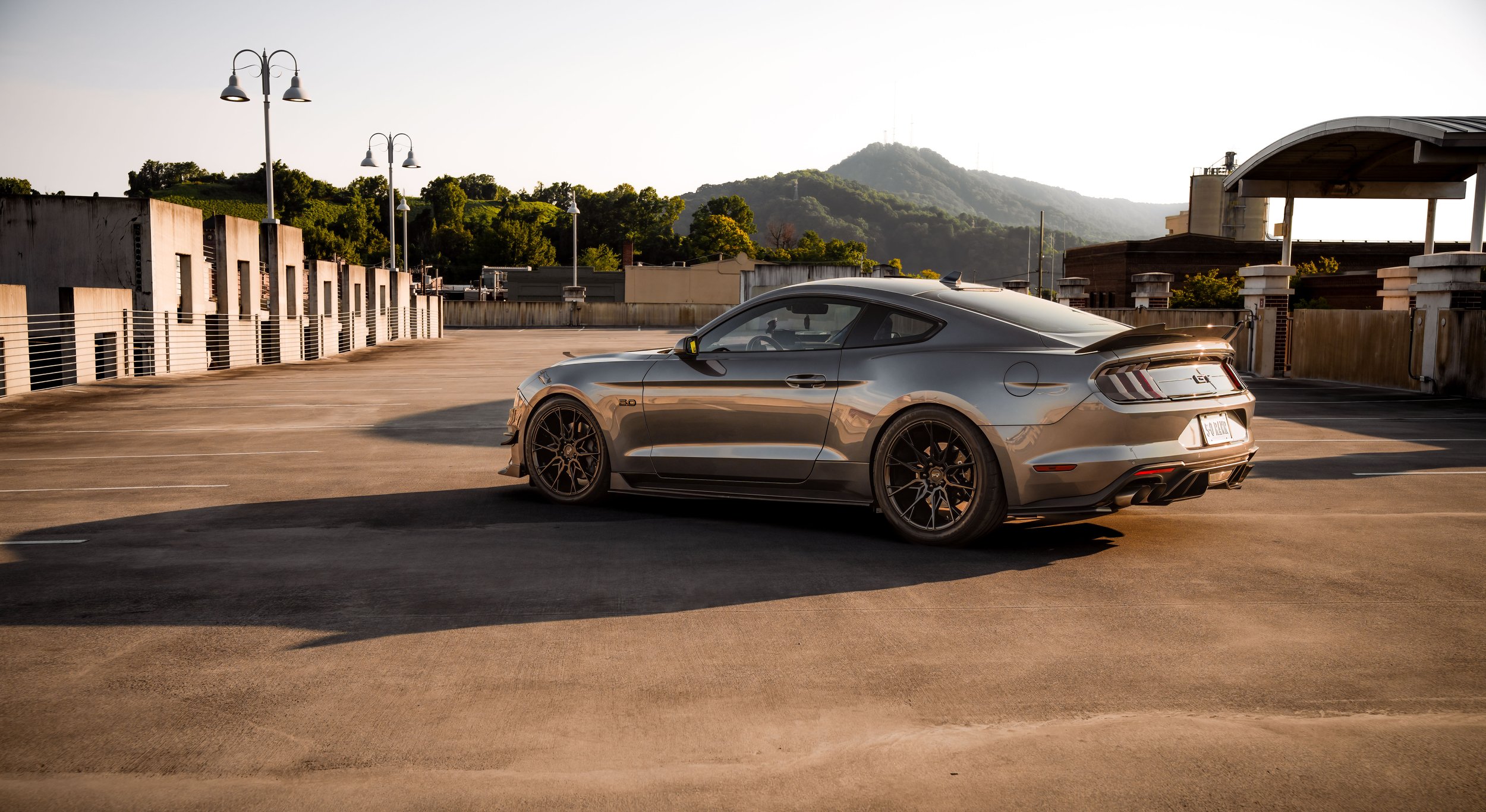 A Gray Ford Mustang GT parked on an outdoor rooftop parking lot during sunset with mountains in the background.