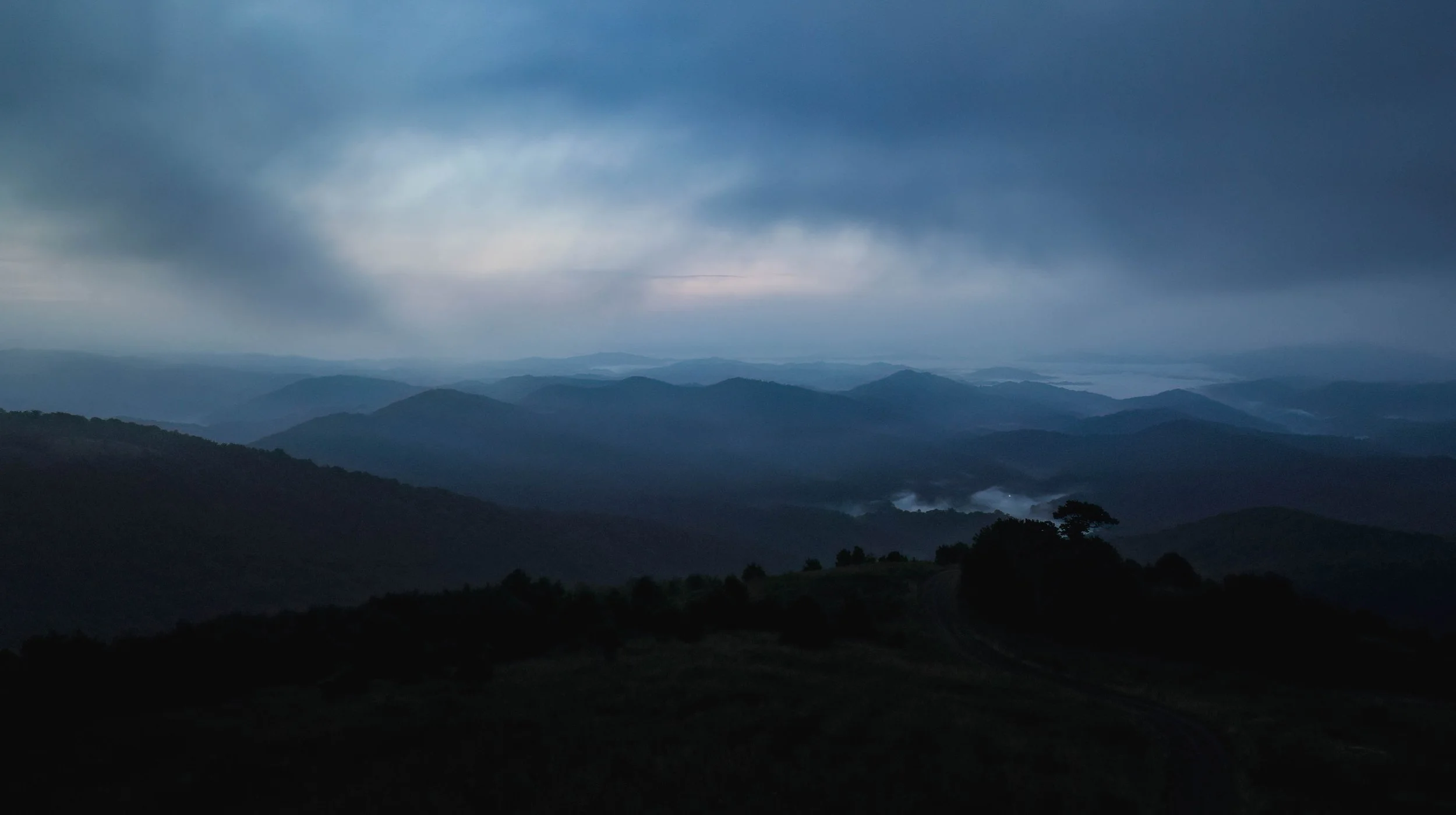 A landscape view of rolling mountains under a dark, cloudy sky with low fog in the valleys.