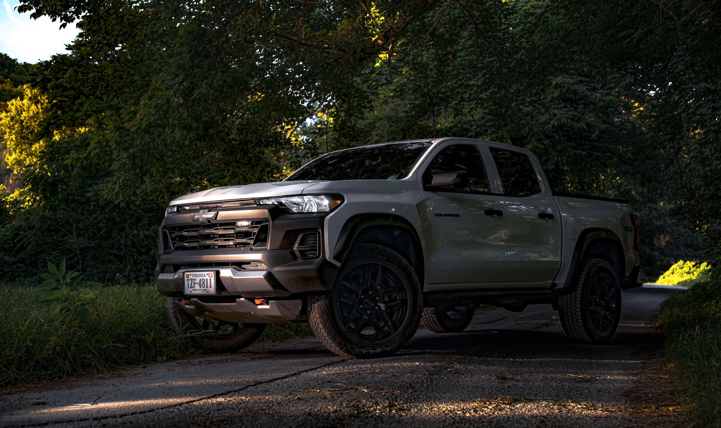A White Chevrolet Colorado pickup truck parked on a gravel road among trees and foliage, with sunlight filtering through the branches.