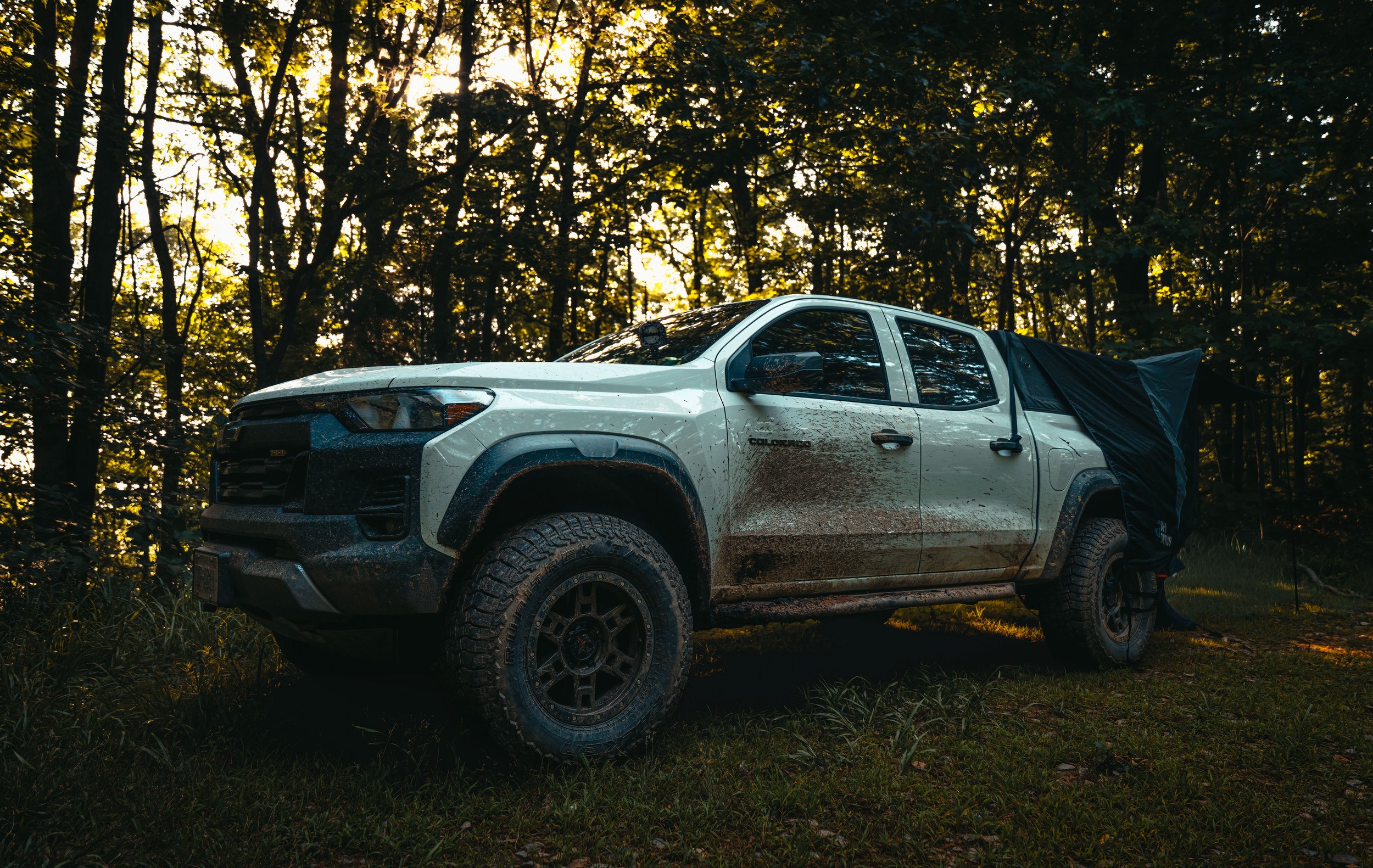 White Chevrolet Colorado Trail Boss pickup truck with muddy tires parked in a forested area during sunset, with tall trees and sunlight filtering through the leaves.