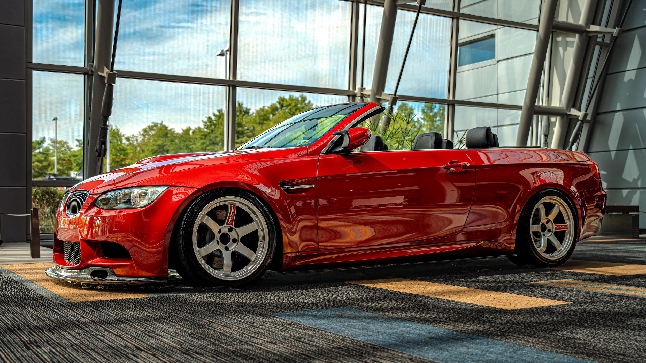 A red convertible sports car parked indoors near large glass windows with a view of trees outside.