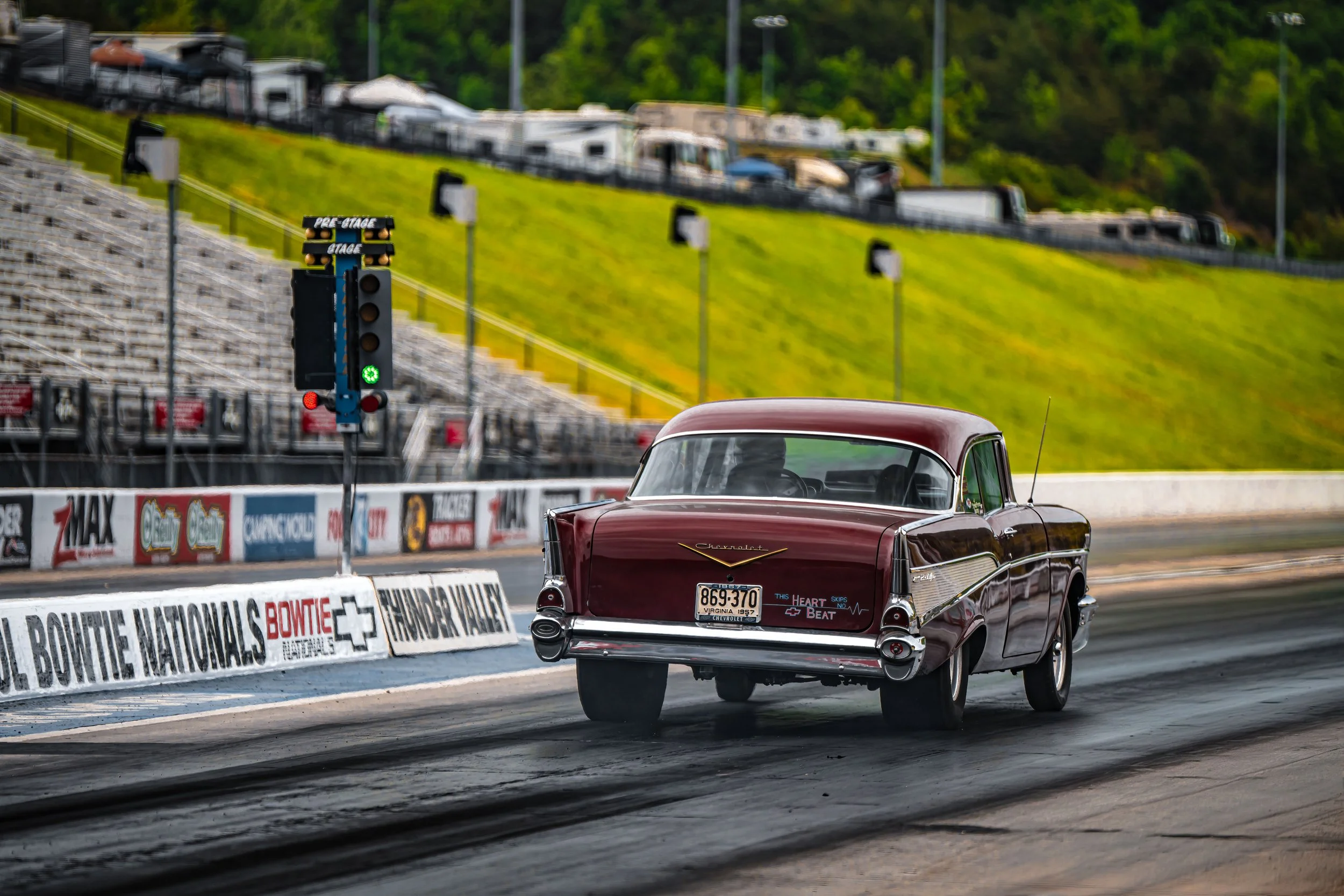A vintage red and black Chevrolet car on a drag strip with a drag racing starting light and empty stands in the background.