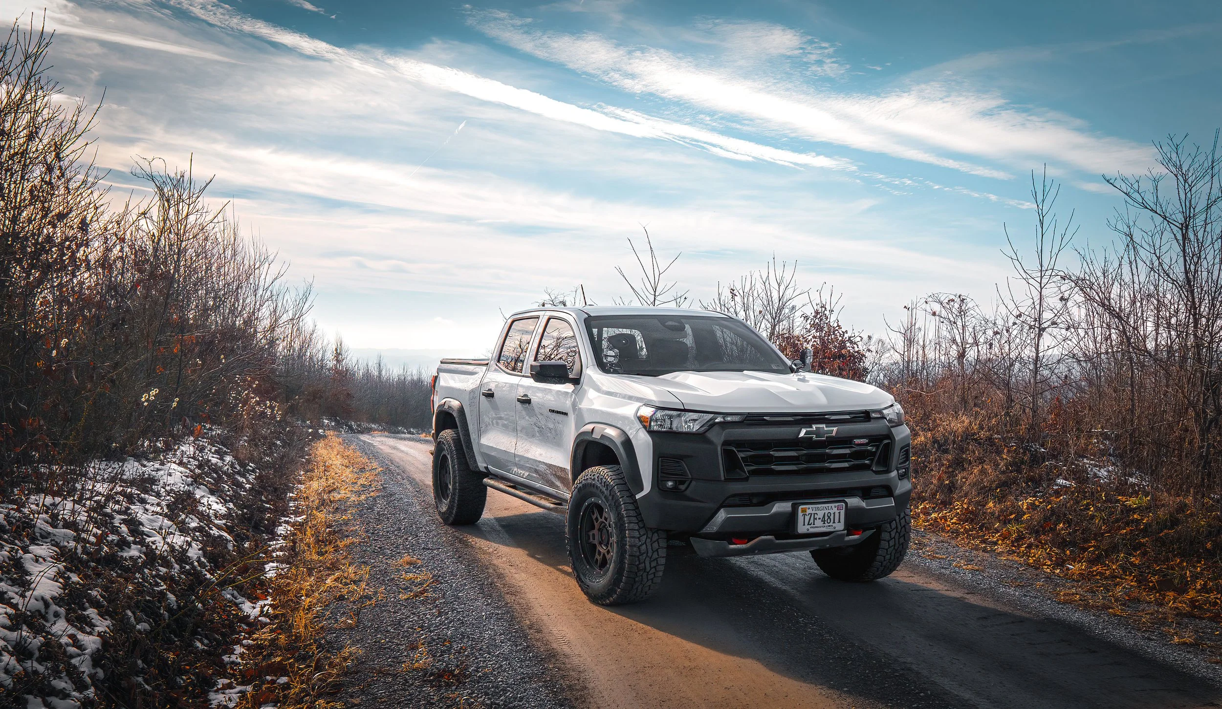 A white Chevrolet pickup truck driving on a dirt and gravel road through a wooded area with snow patches, under a blue sky with wispy clouds.
