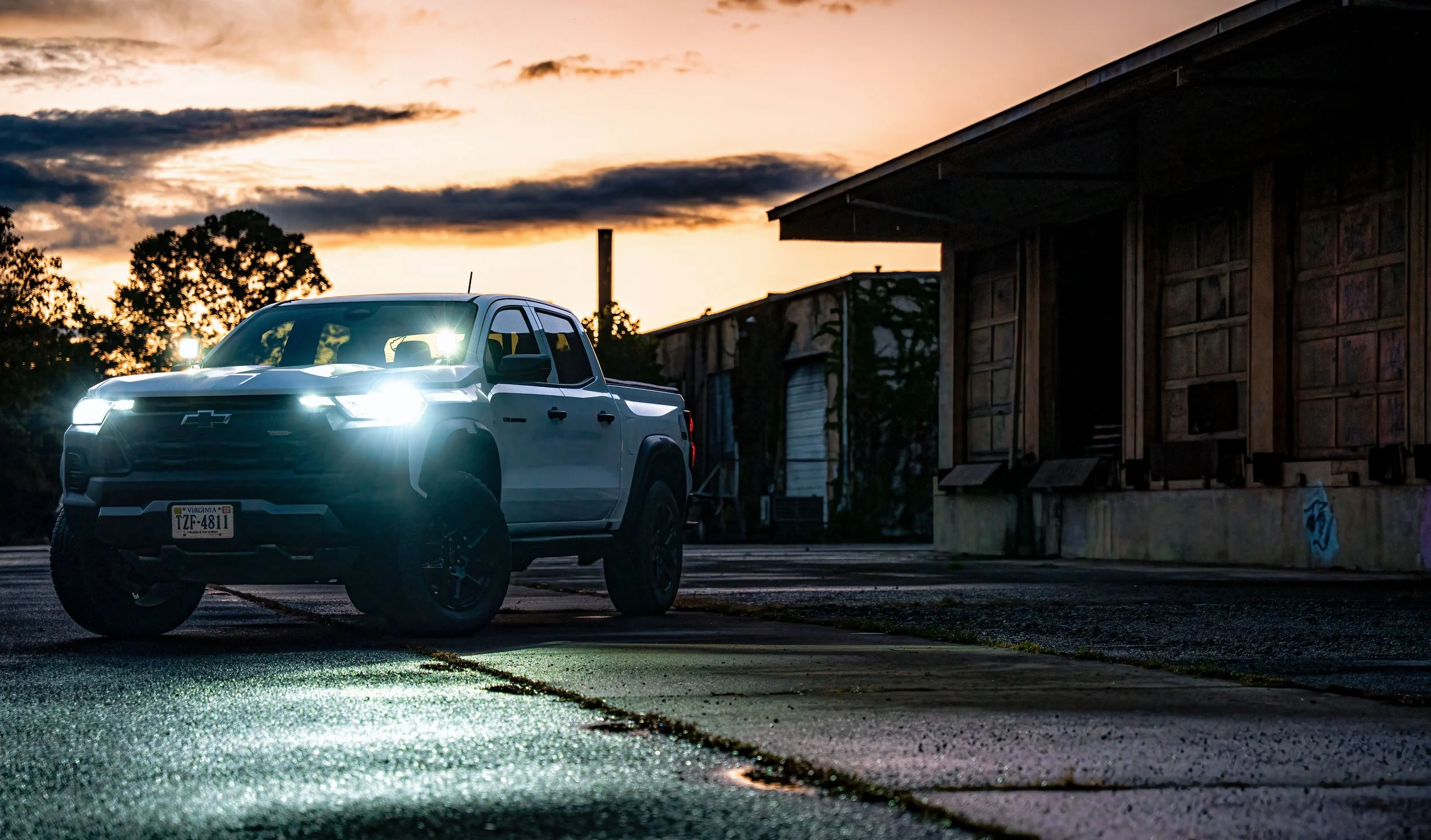 A white Chevrolet pickup truck with its headlights on parked on a wet surface during sunset, next to an abandoned building.