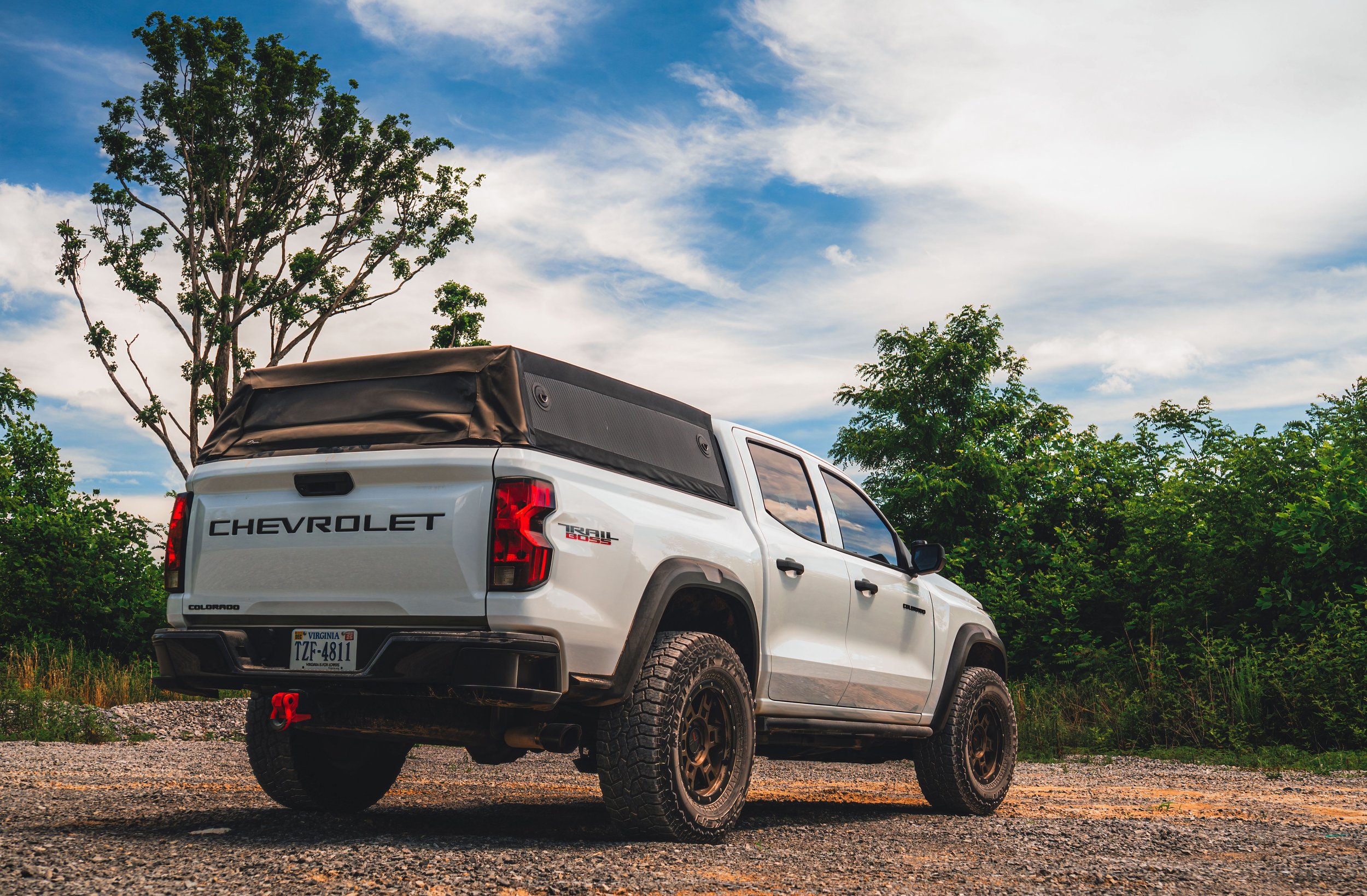 A white Chevrolet Colorado pickup truck with a black cover over the bed, parked outdoors on a dirt and gravel surface, with green trees and a partly cloudy sky in the background.