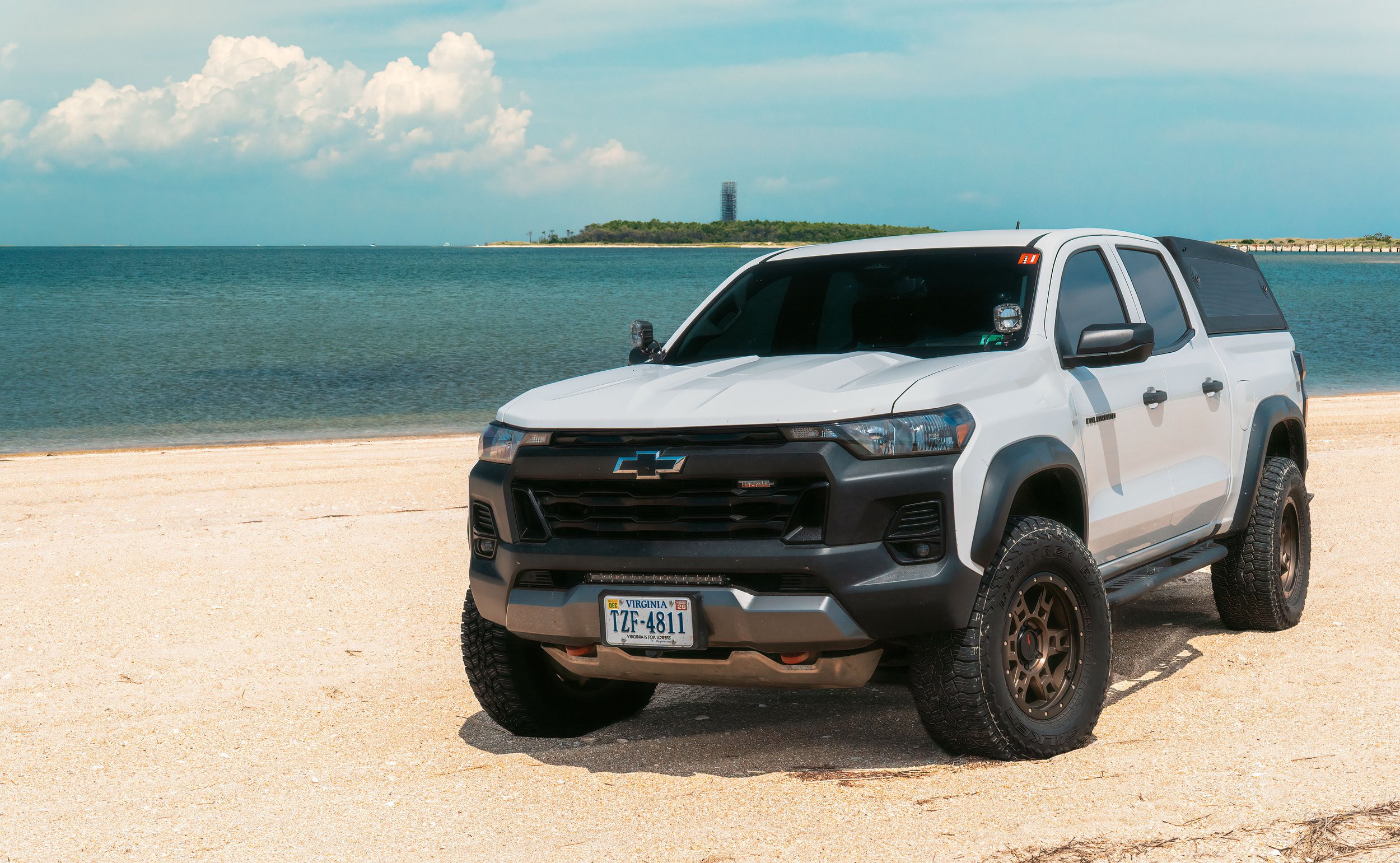A white Chevrolet Colorado pickup truck parked on a sandy beach near the water with an island and cloudy sky in the background.