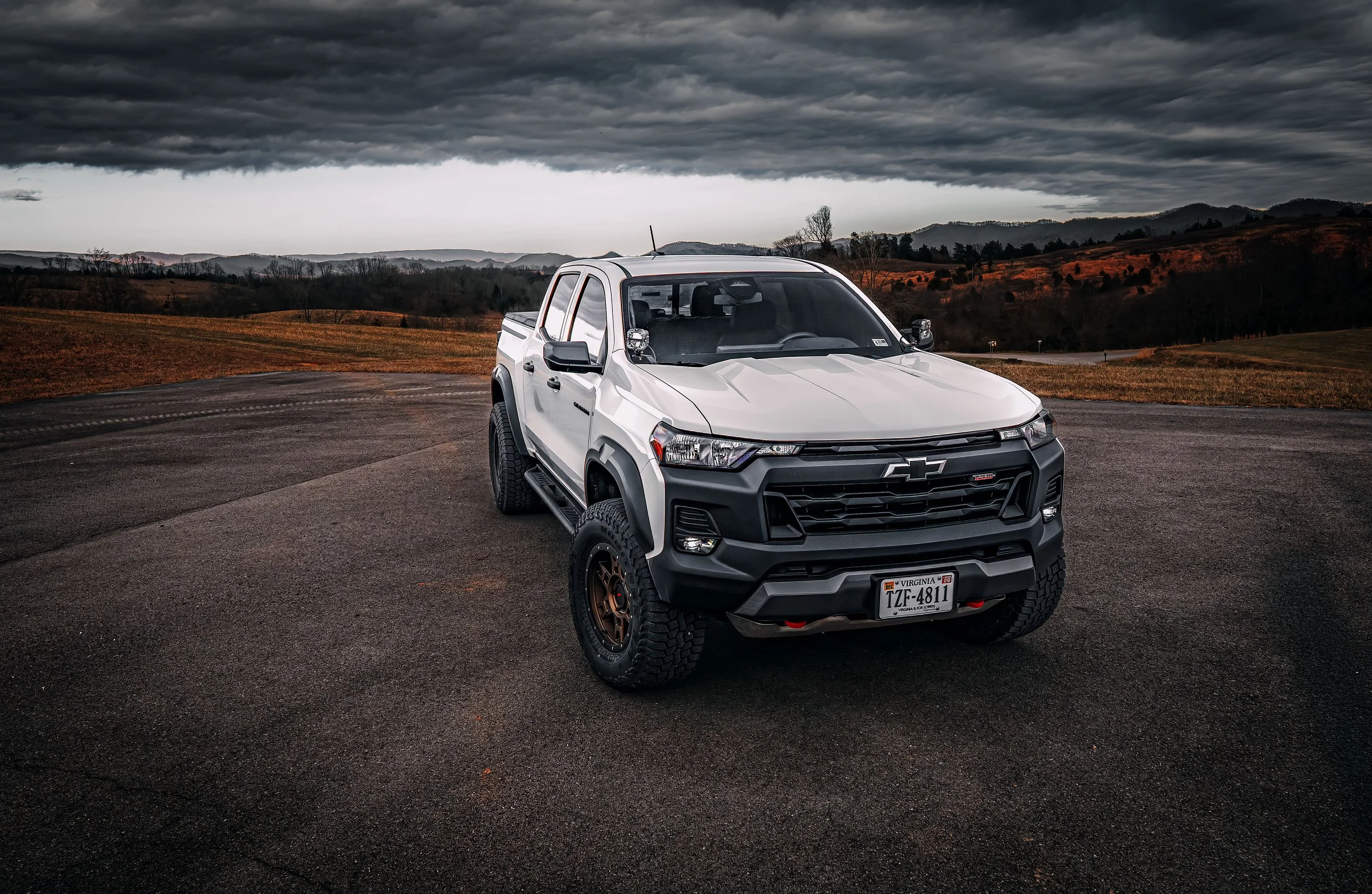 A white Chevrolet pickup truck parked on a dark asphalt road with a mountainous landscape and stormy cloudy sky in the background.