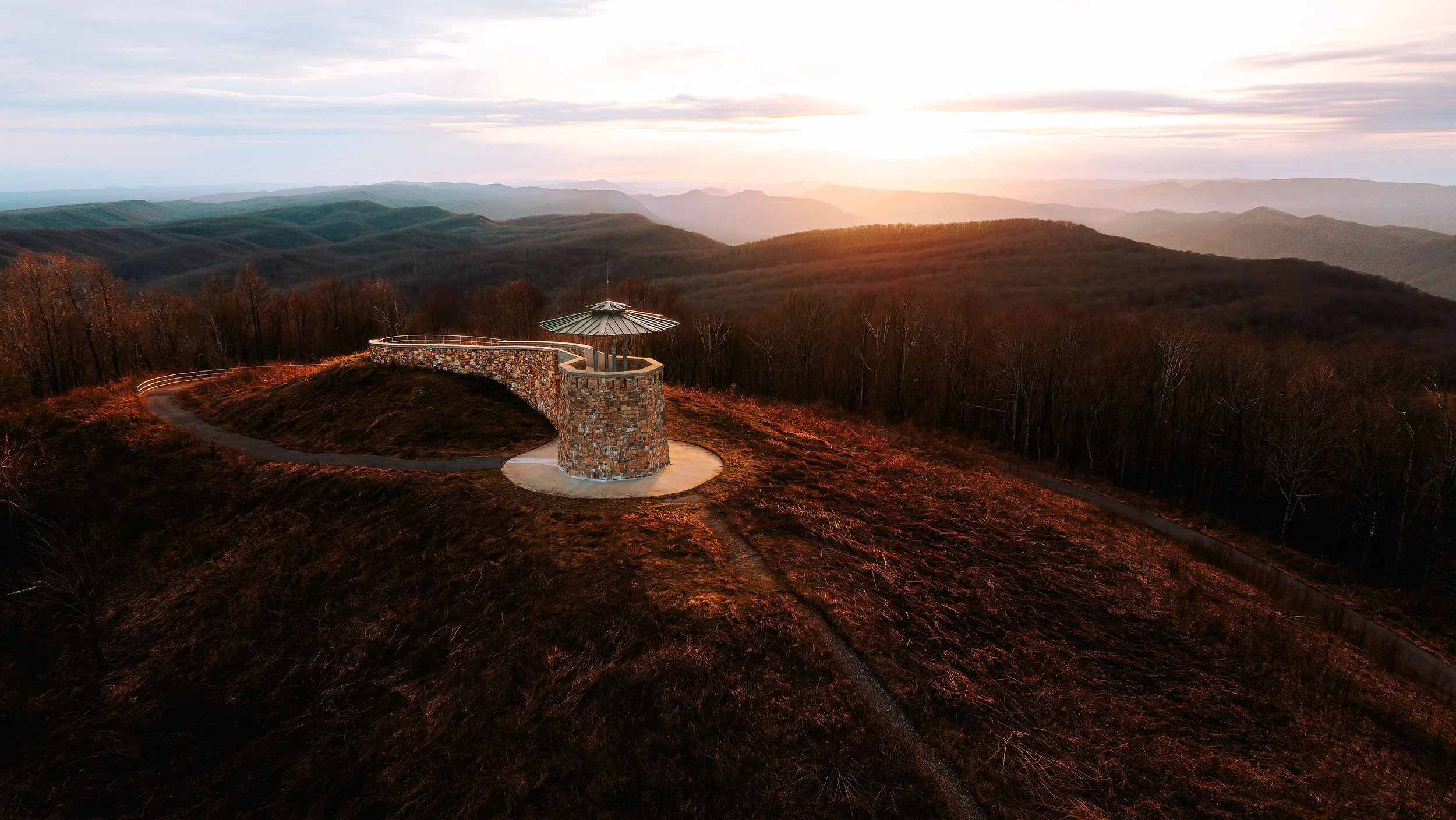 Scenic view of mountain ranges at sunset with a stone observation tower and a winding pathway in the foreground.