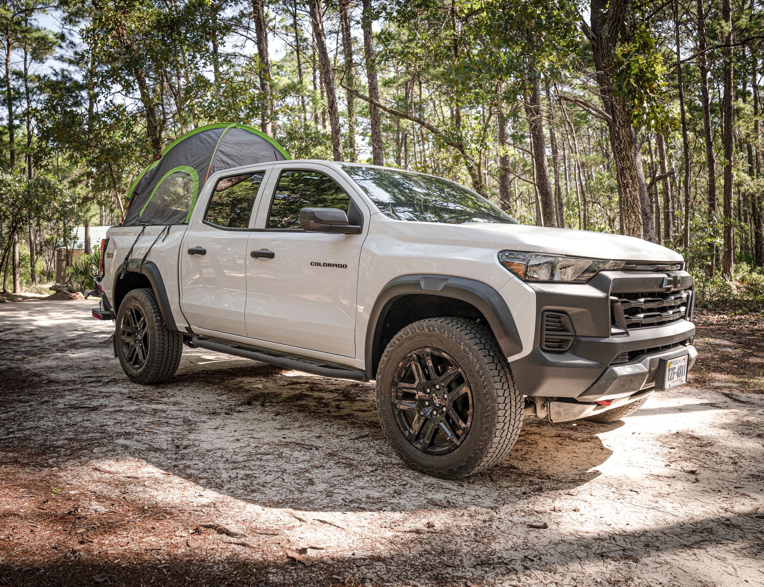 White Chevrolet Colorado pickup truck with a camping tent attached to the back, parked in a wooded area with trees.