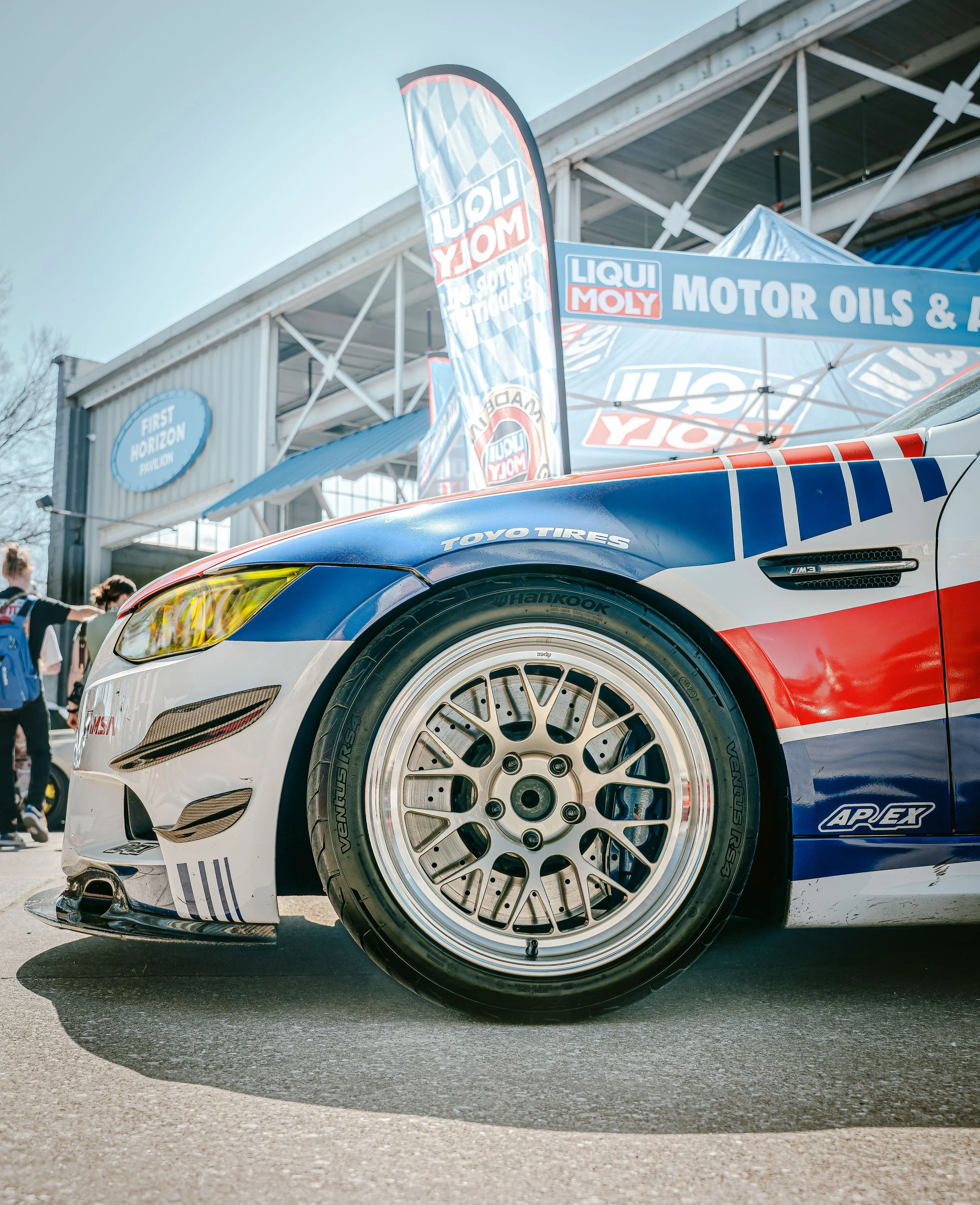 Close-up of a race car's front wheel and side, with tire brands Toyo Tires and Hankook visible, at a motorsport event with banners and people in the background.