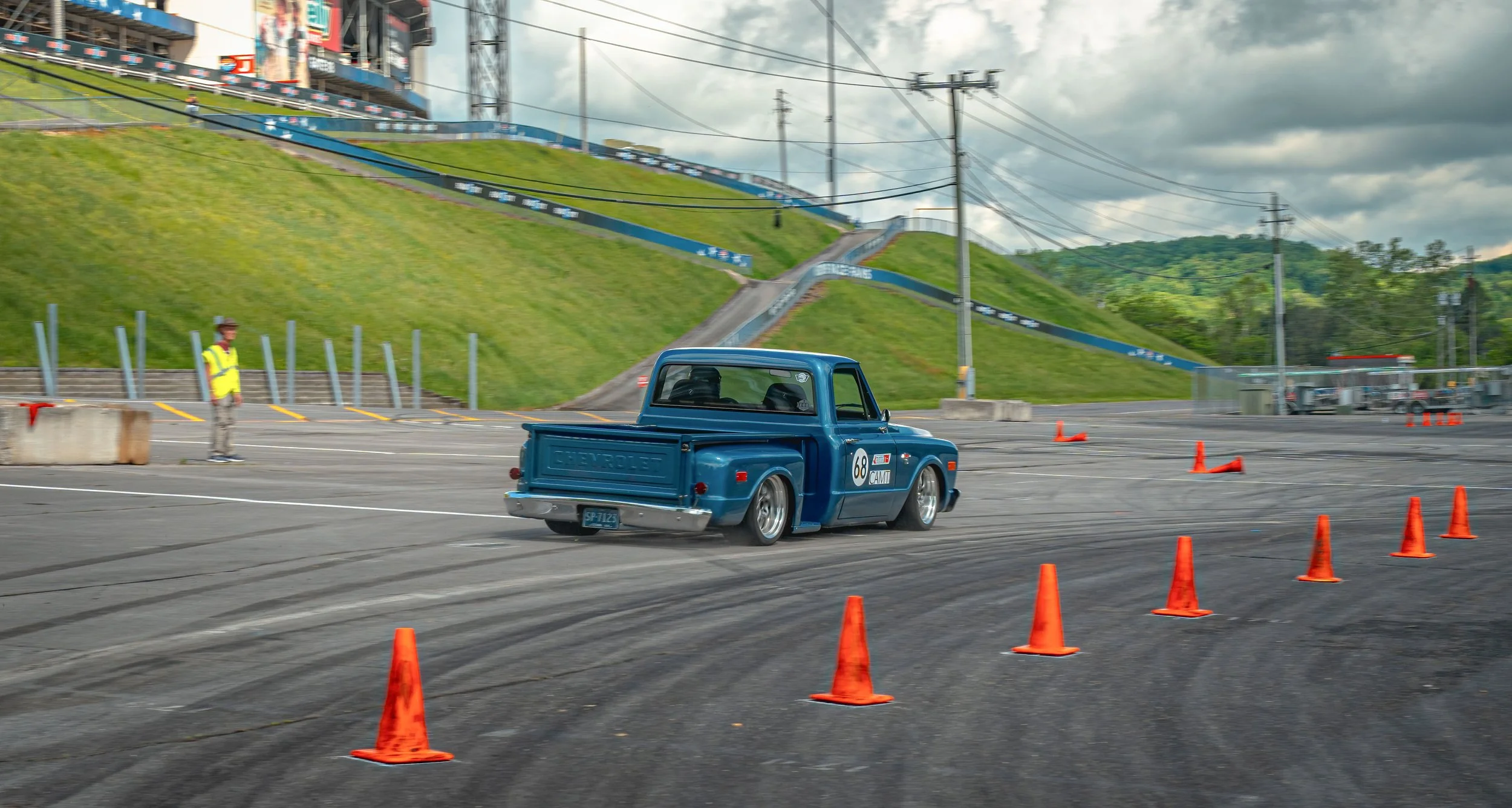 A vintage blue Chevrolet pickup truck participating in a car rally, driving through a course marked with orange cones, with a man in a yellow safety vest standing nearby, in an open parking lot.