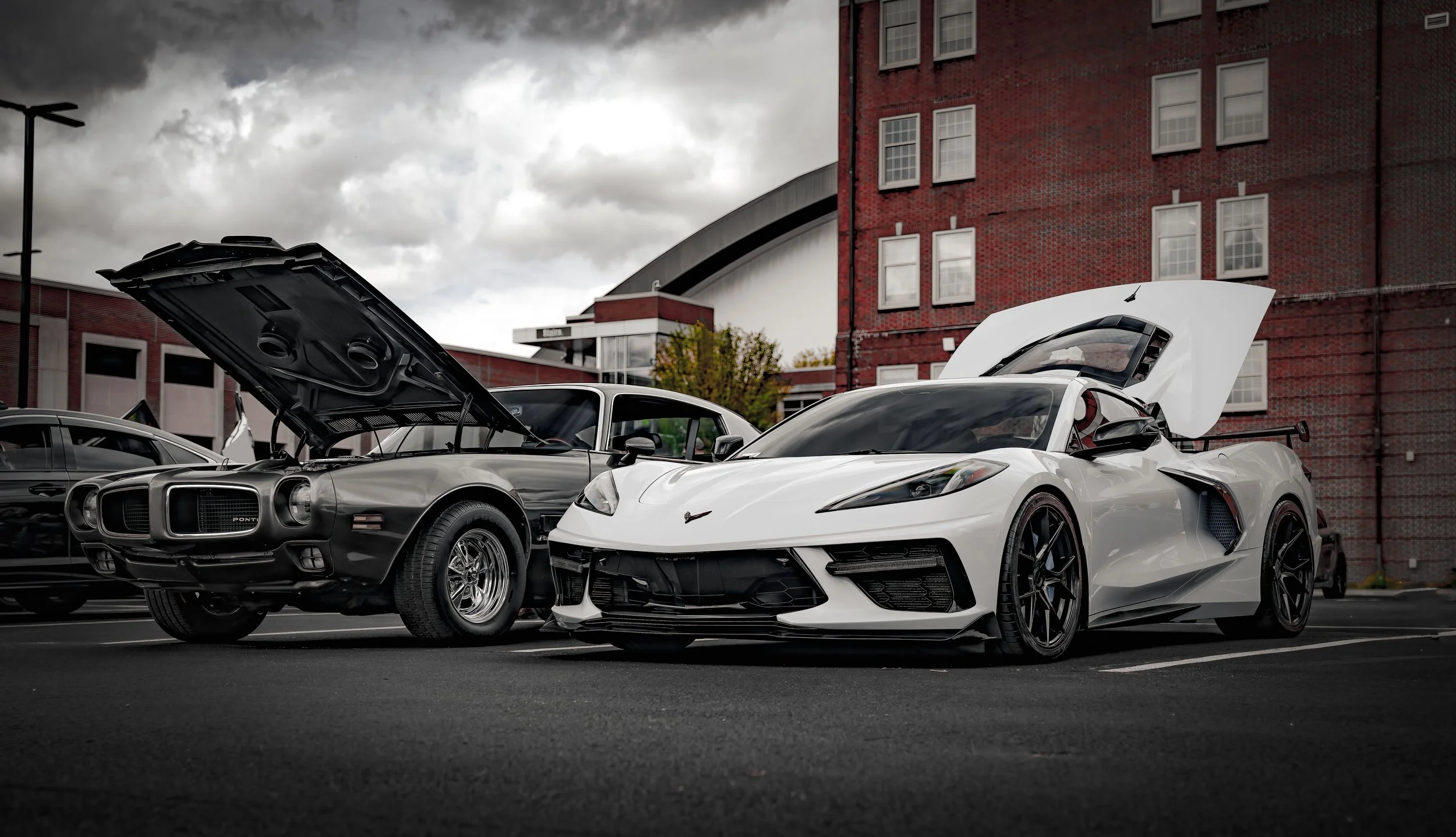 A black vintage Pontiac muscle car and a white modern sports car with gullwing doors open, parked outdoors near a red brick building under a cloudy sky.