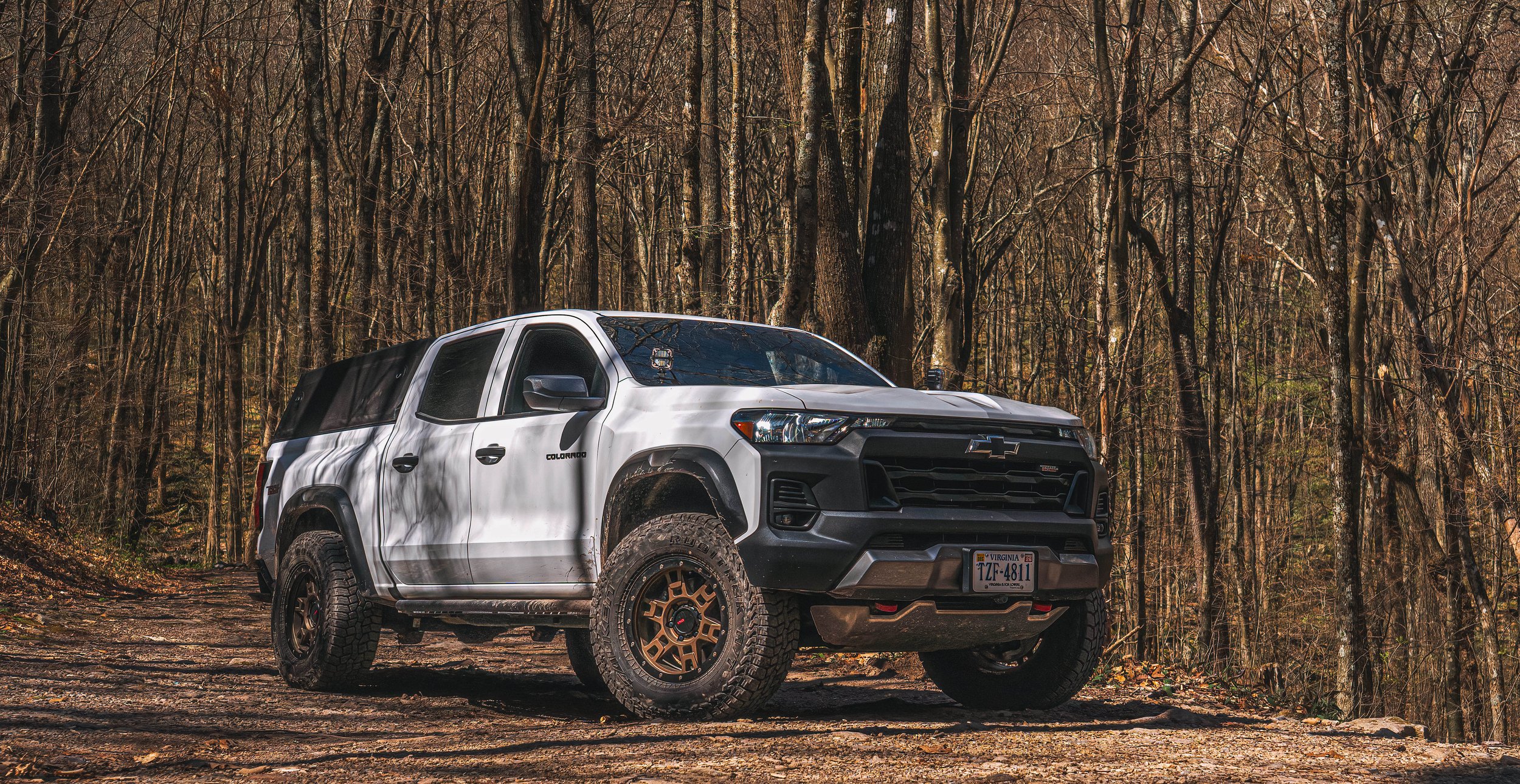A white Chevrolet Colorado pickup truck parked on a dirt path in a dense forest with leafless trees.