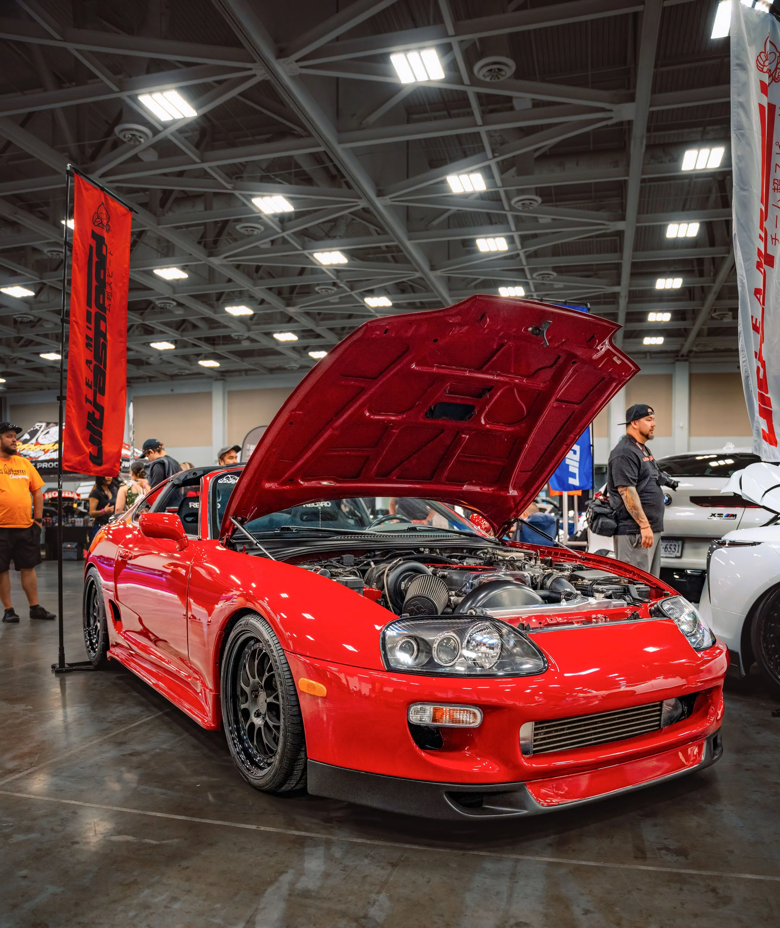 Red sports car with hood open on display at an indoor car show, surrounded by people and banners.