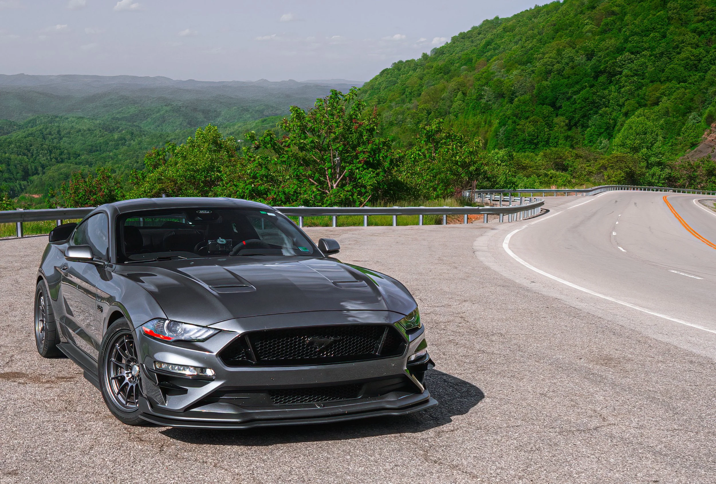 A gray Ford Mustang sports car parked on the side of a winding mountain road surrounded by green trees and hills.