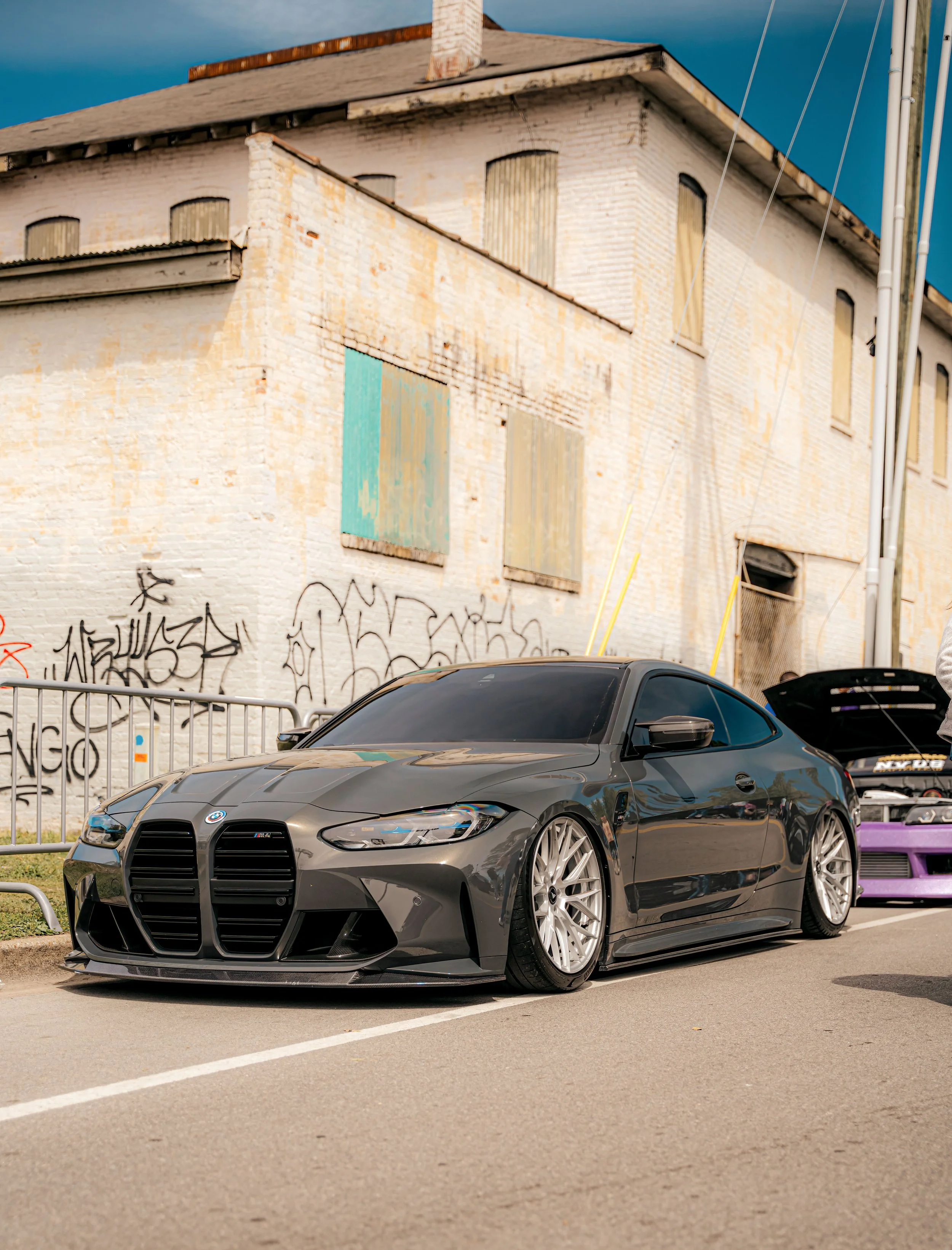 A sleek, dark gray BMW M4 sports car parked on a street next to an old brick building with graffiti, with another car visible in the background with its hood open.