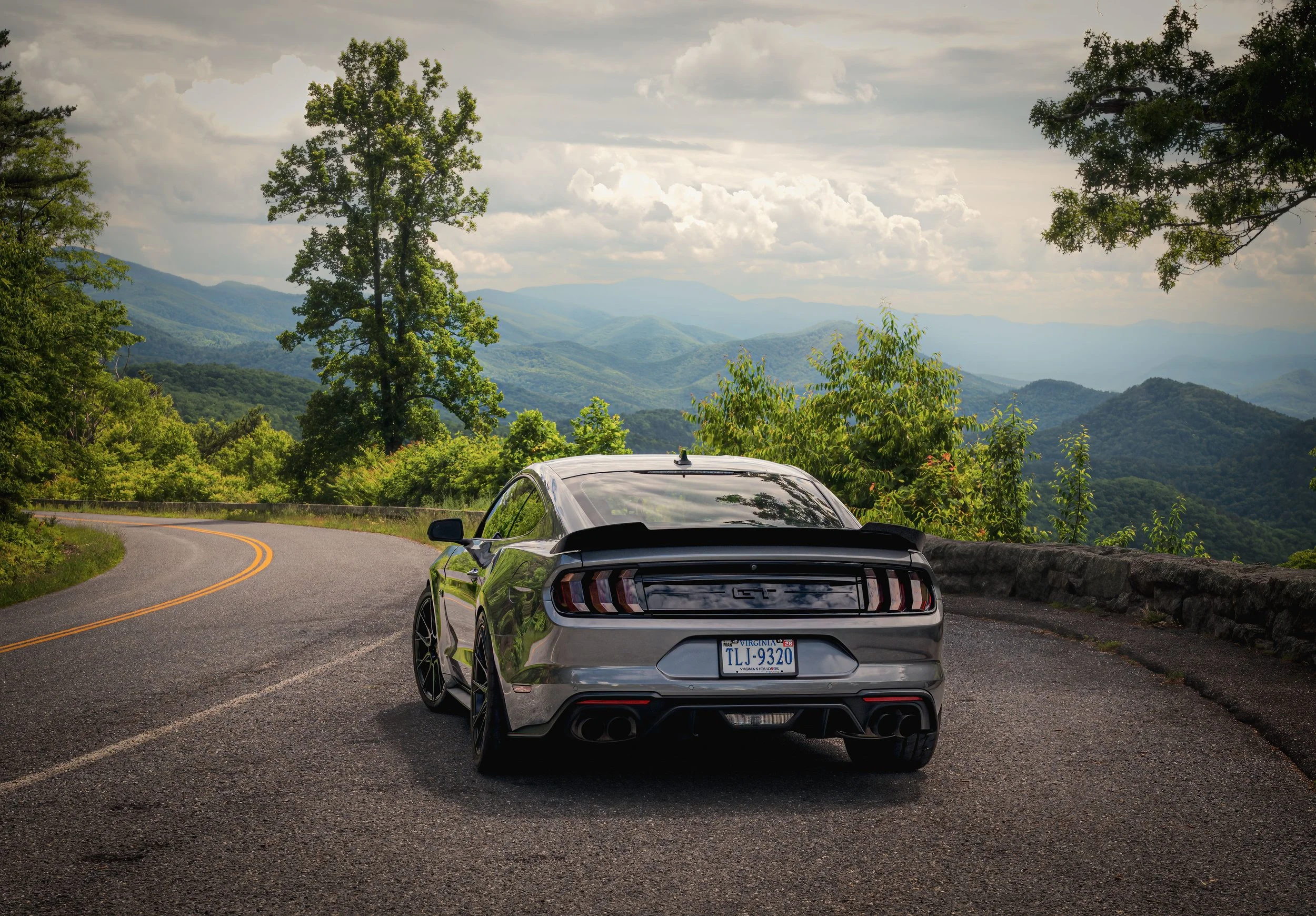 A silver sports car parked on a mountain road with lush green trees and rolling hills in the background.