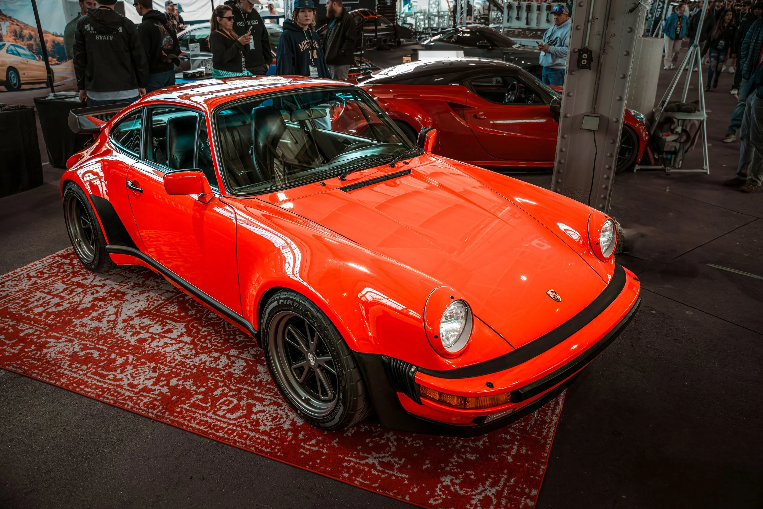 A red classic Porsche sports car displayed on a decorative rug at an indoor car show, with people looking and taking photos in the background.