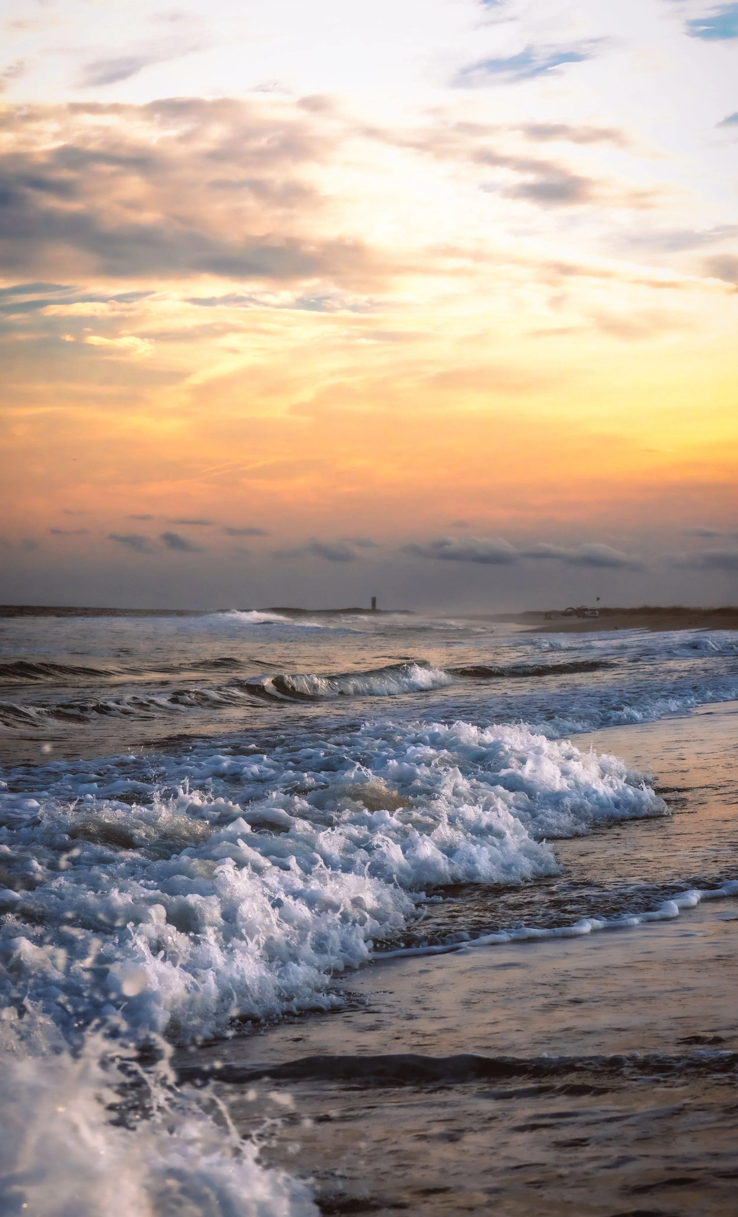 Sunset over ocean with clouds and waves crashing on shore.