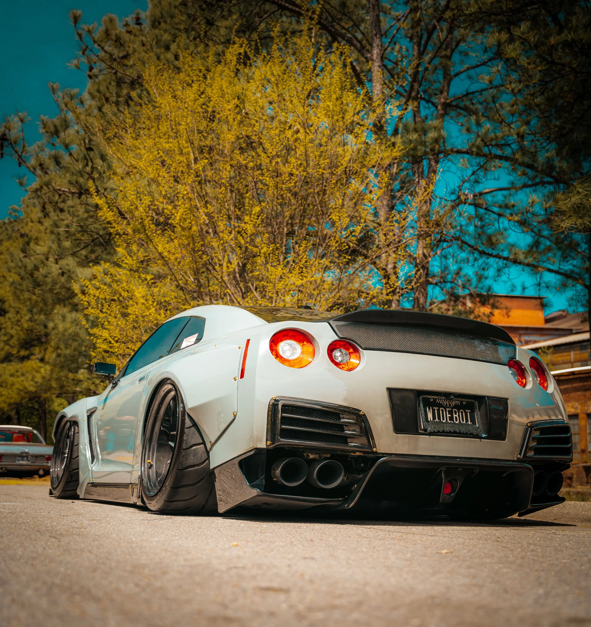 A customized white Nissan R35 GTR with modified body kit and wide tires parked on a street at night, with trees and buildings in the background.