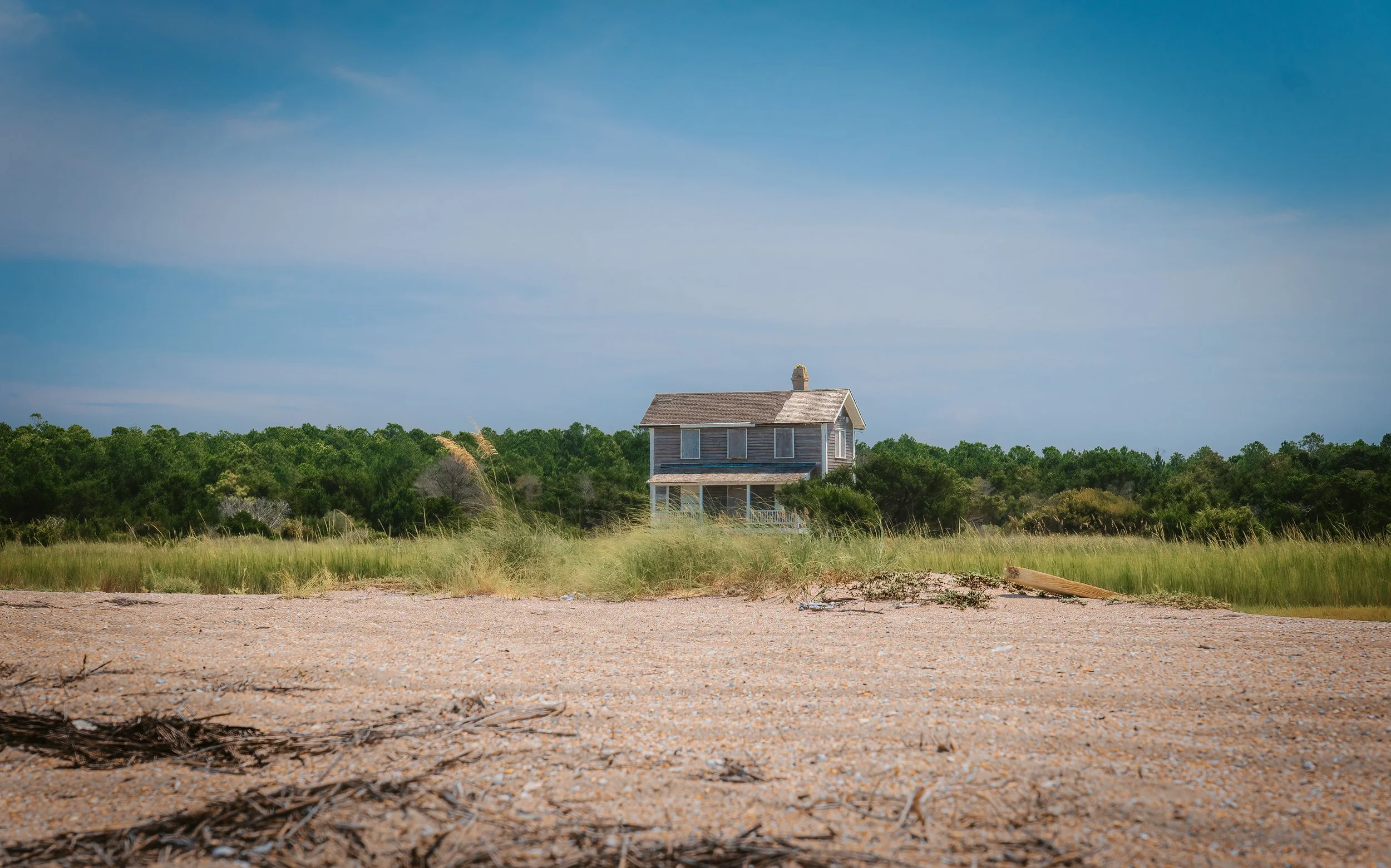 A wooden house in a grassy field with a green forest background under a blue sky. Cape Lookout, NC 