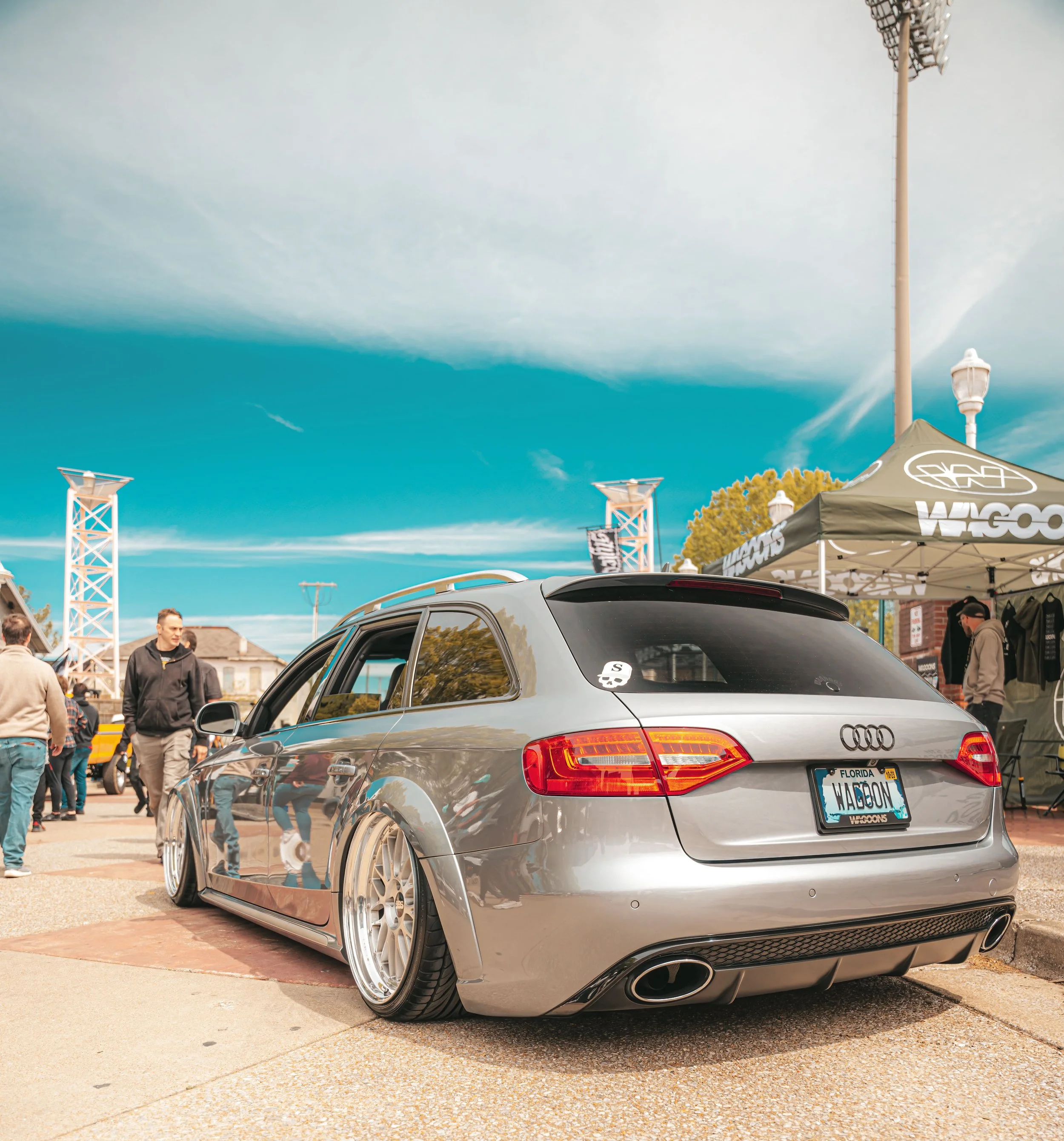 Silver Audi station wagon with custom wheels parked at an outdoor car show, with people and tents in the background.